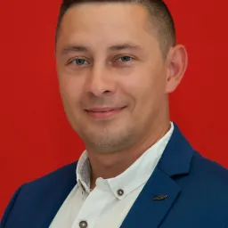 Portret uśmiechniętego mężczyzny w niebieskiej marynarce i białej koszuli na czerwonym tle, z przypinką w kształcie pióra na klapie marynarki.