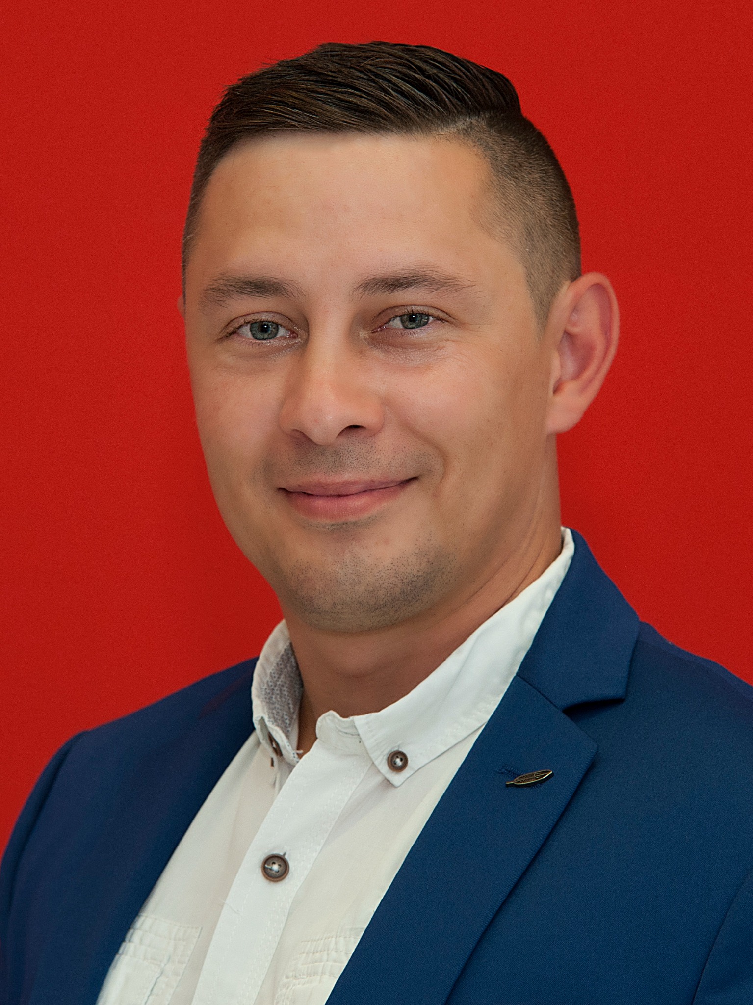 Portret uśmiechniętego mężczyzny w niebieskiej marynarce i białej koszuli na czerwonym tle, z przypinką w kształcie pióra na klapie marynarki.