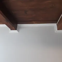 Drewniany strop z belkami i białą sztukaterią przy szarej ścianie, widok z dołu.