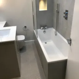 Nowoczesna łazienka z wanną, szarą glazurą, wnęką z oświetleniem, umywalką z białym blatem i toaletą w szaro-białej kolorystyce.