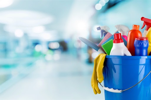 Niebieskie wiadro pełne środków czystości, gąbek i żółtych rękawic na rozmytym tle korytarza.