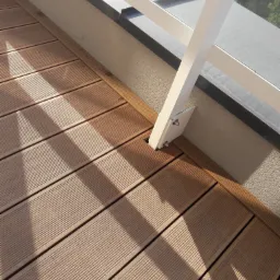 Nowoczesny balkon z jasnobrązową podłogą z desek, biała balustrada z widocznymi mocowaniami, cień rzucany przez konstrukcję na podłogę.