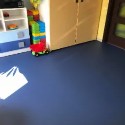 Niebieska podłoga z wykładziny w pokoju dziecięcym, zabawki przy ścianie, szafki z jasnego drewna i drzwi w tle.