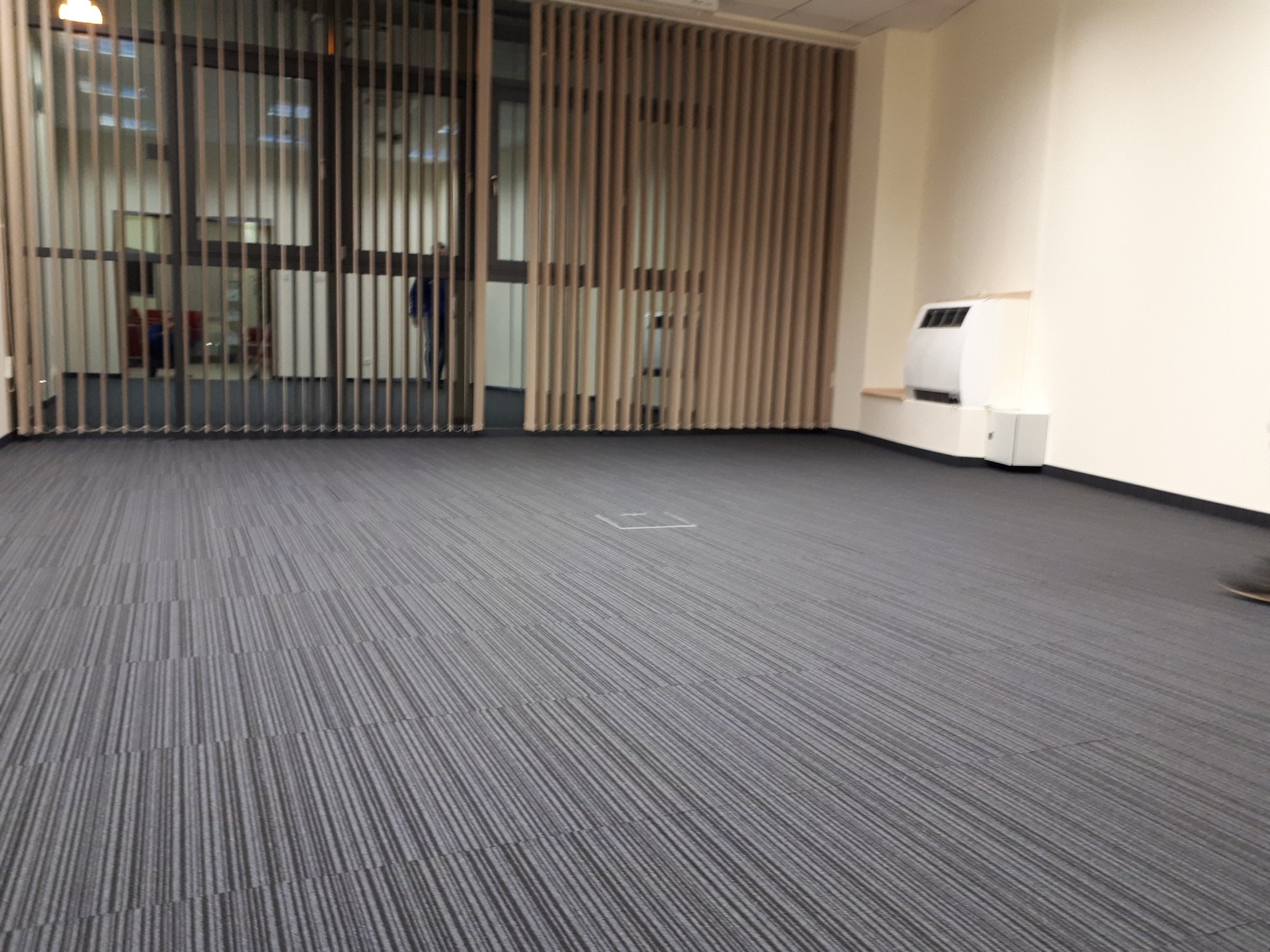 Szeroki kadr na szarą wykładzinę dywanową w biurze, widoczne pionowe żaluzje okienne i biały grzejnik elektryczny przy ścianie.