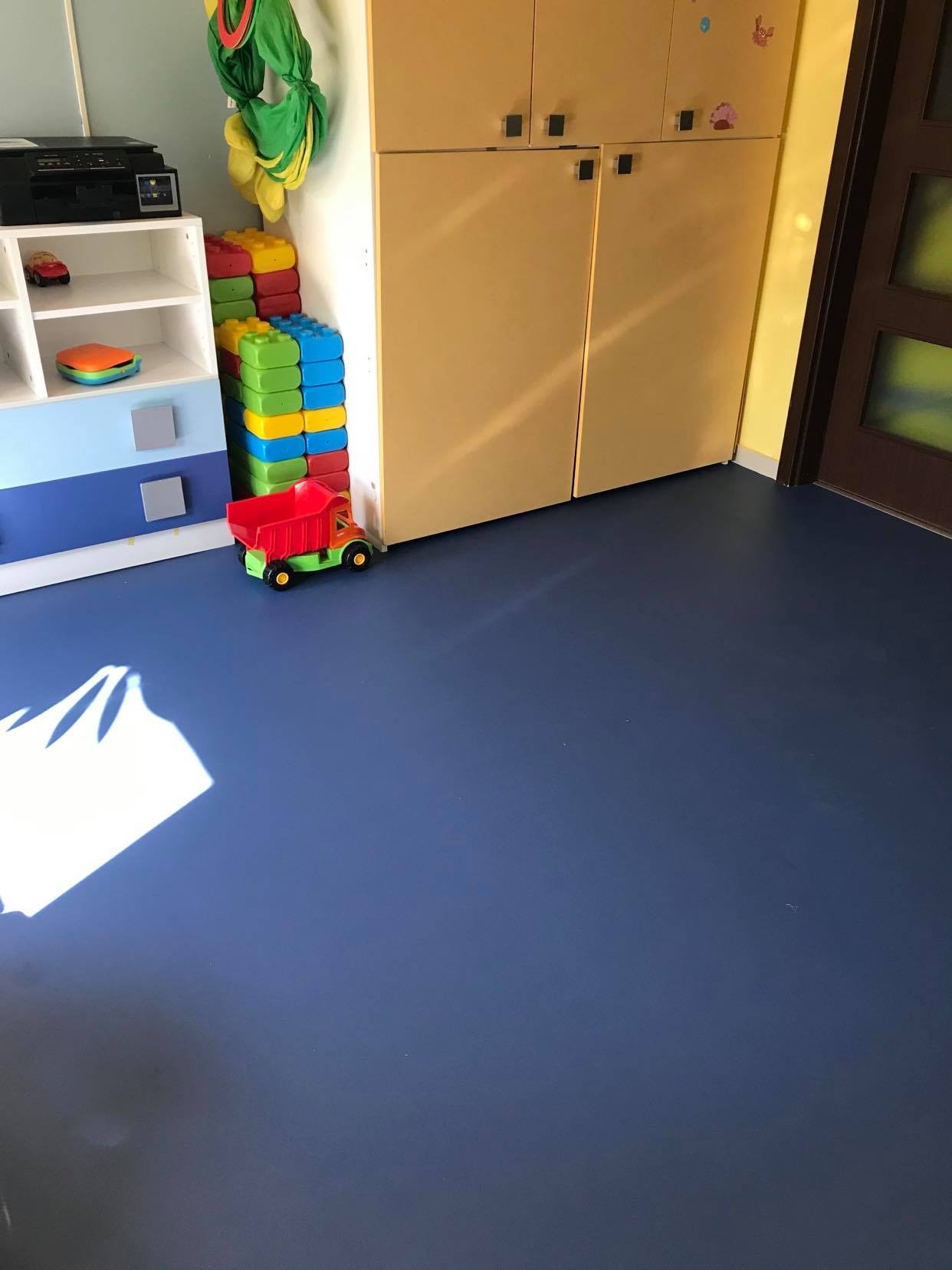 Niebieska podłoga z wykładziny w pokoju dziecięcym, zabawki przy ścianie, szafki z jasnego drewna i drzwi w tle.