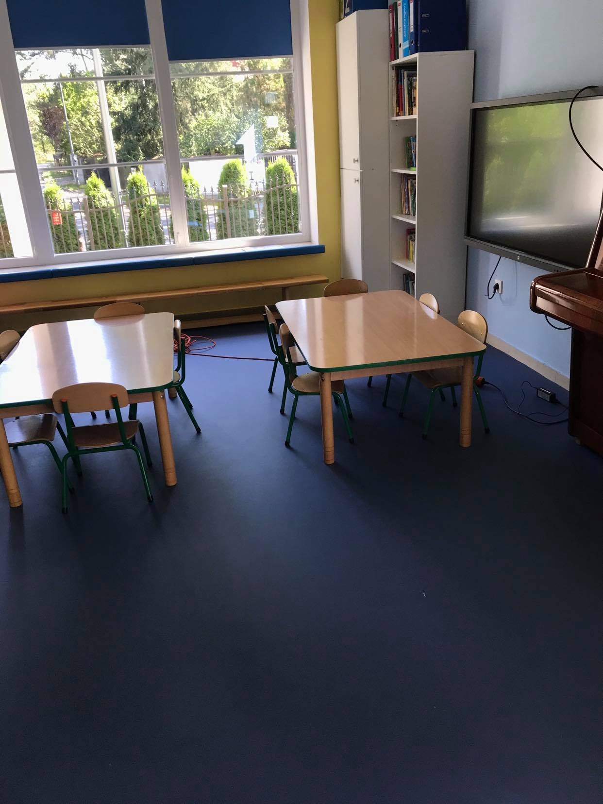 Klasa przedszkolna z ciemnoniebieską podłogą, drewnianymi stolikami i krzesłami, regałem z książkami i interaktywną tablicą.