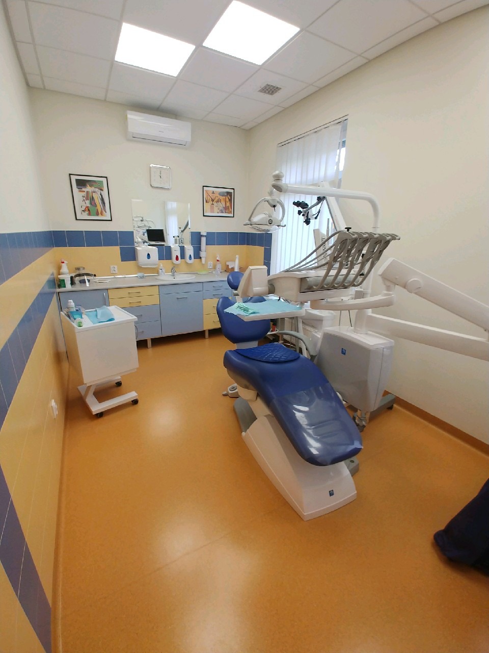 Wnętrze gabinetu dentystycznego z niebieskim fotelem, żółtą podłogą i szafkami w odcieniach niebieskiego i beżu, widok na umywalkę i sprzęt medyczny. Na ścianach wiszą obrazy.
