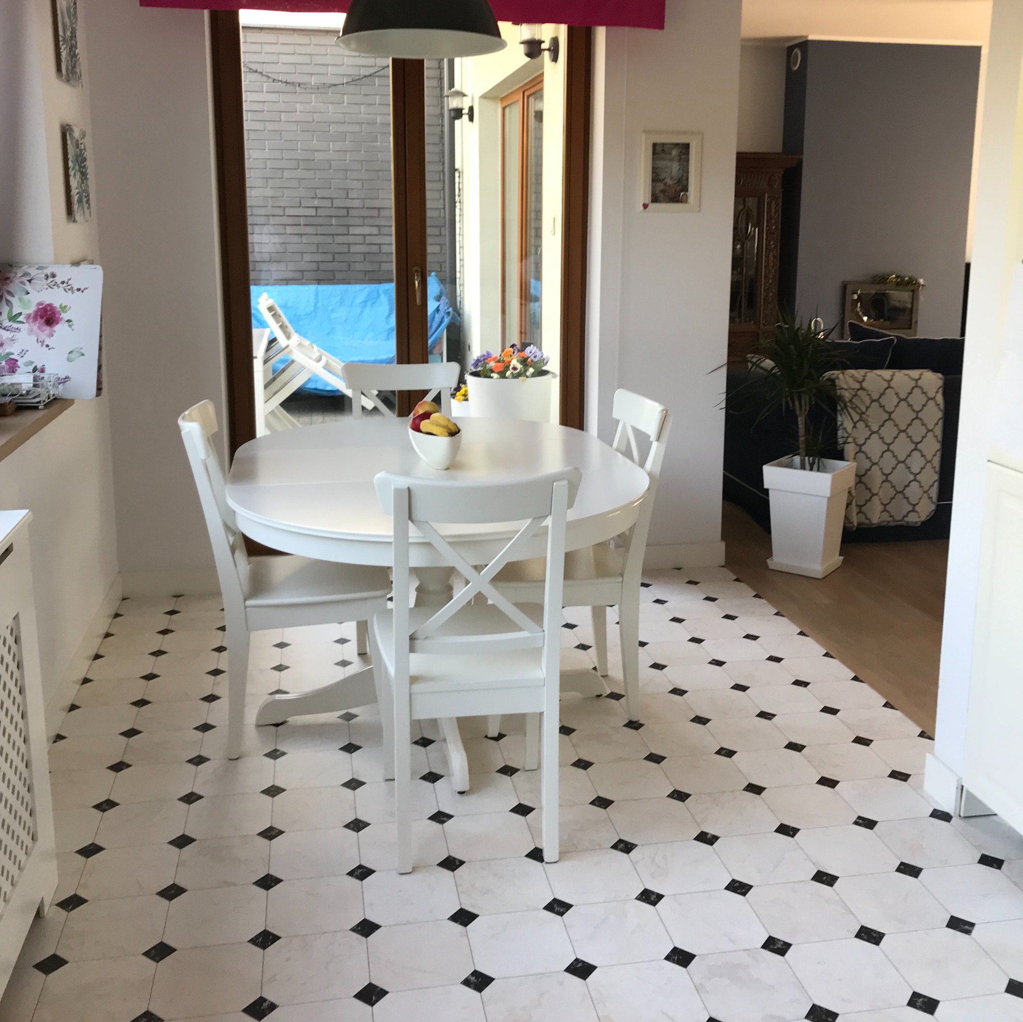 Jasna kuchnia z białym, okrągłym stołem i krzesłami na podłodze z białych płytek z czarnymi akcentami, widok na taras z niebieskim leżakiem.