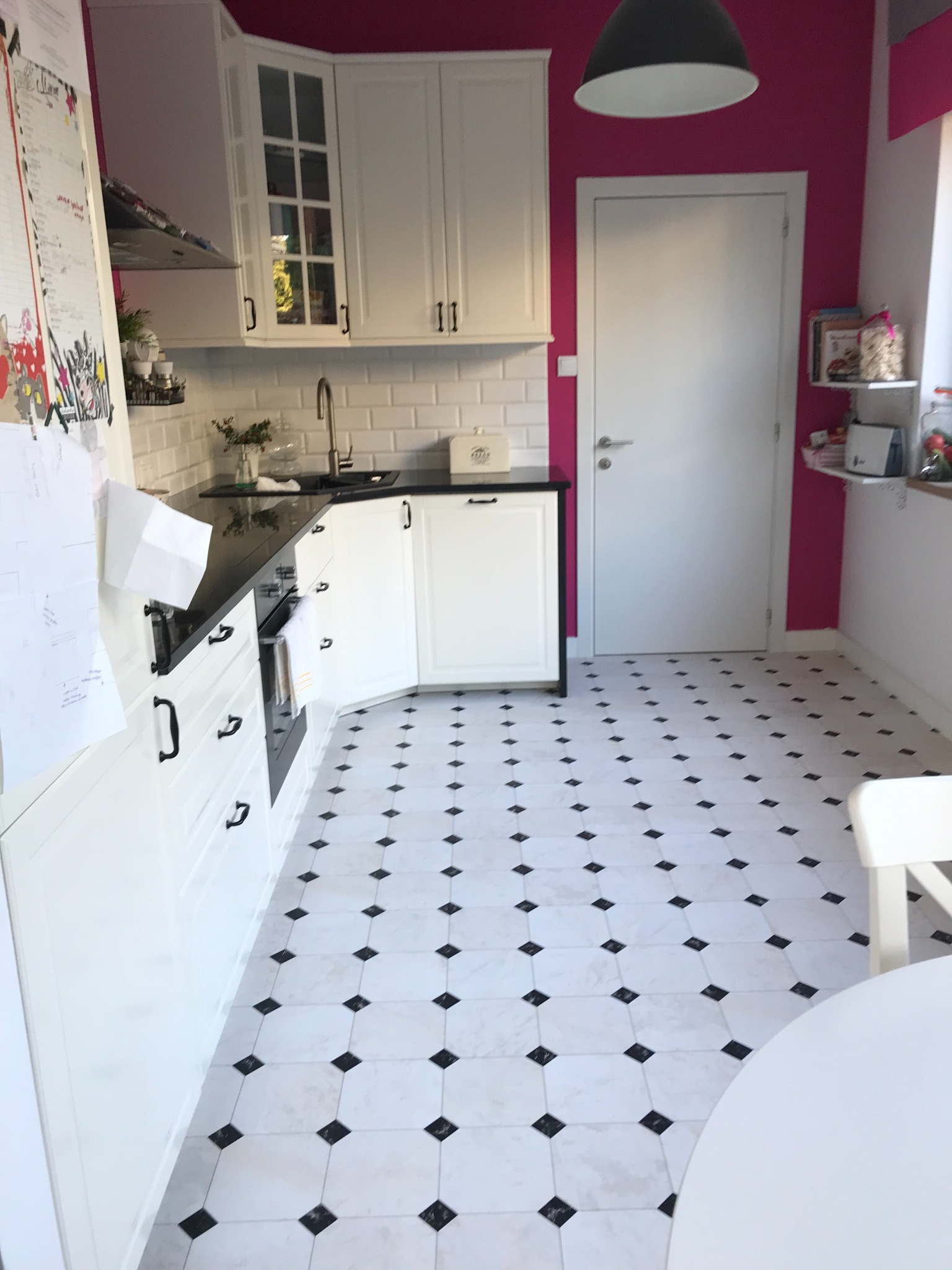 Jasna kuchnia z białymi szafkami, czarnym blatem i podłogą w biało-czarną szachownicę, ściana pomalowana na intensywny różowy kolor.