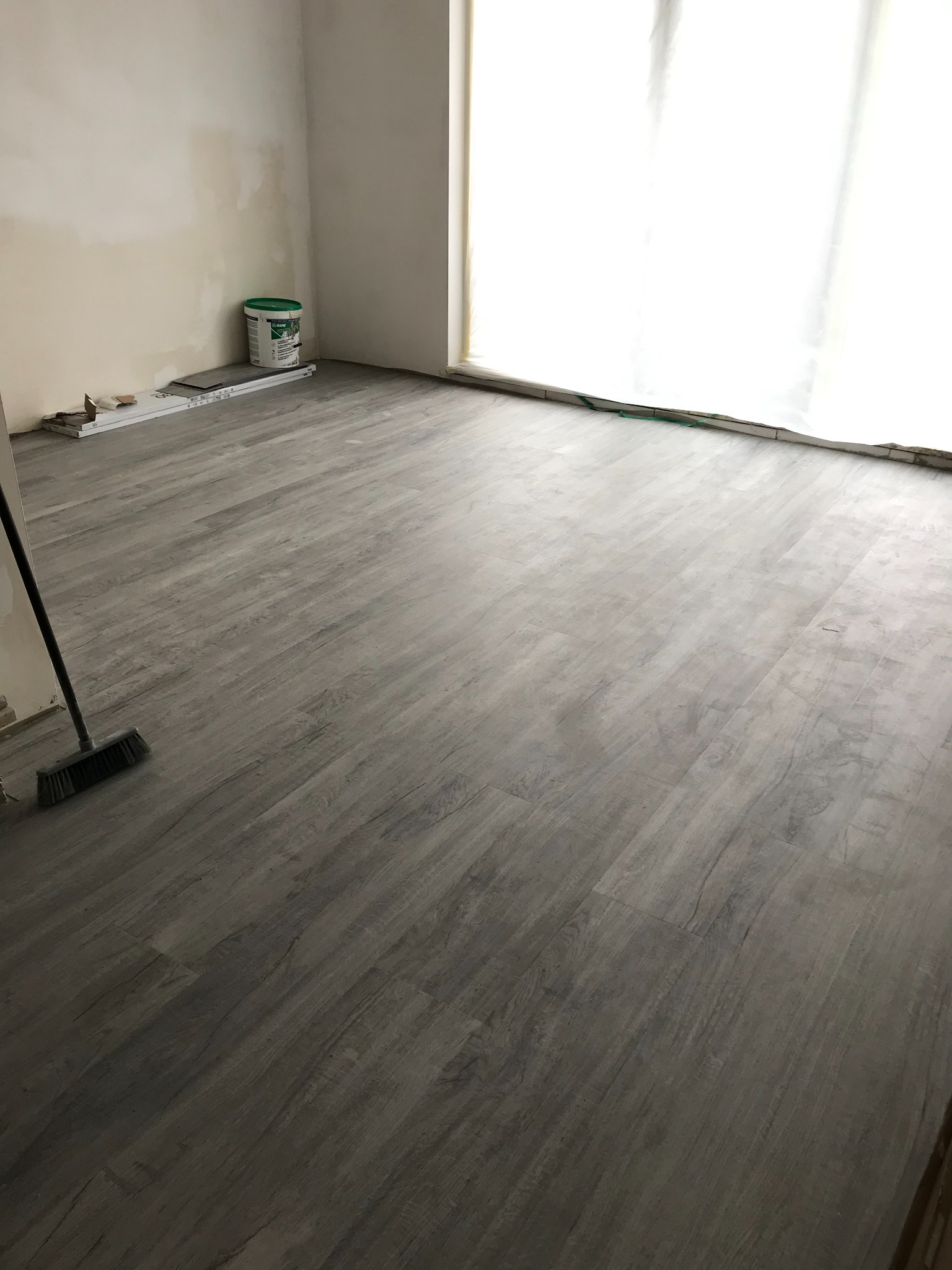 Świeżo ułożone panele podłogowe w odcieniach szarości, widoczna szczotka do zamiatania i wiadro z klejem przy ścianie, przygotowane do dalszych prac wykończeniowych.