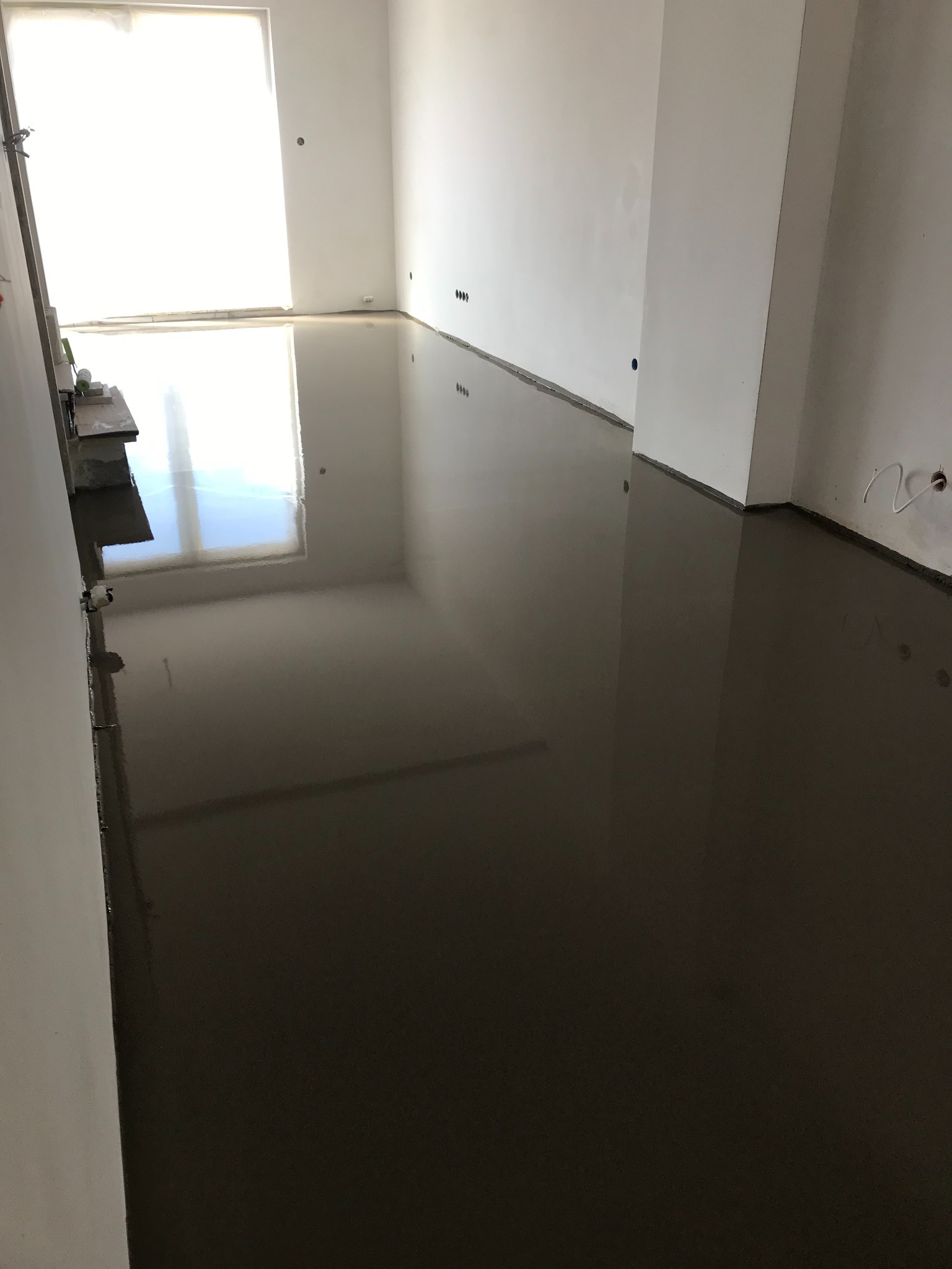 Świeżo wylana, samopoziomująca wylewka podłogowa w jasnym pomieszczeniu z białymi ścianami, widoczne odbicie okna w mokrej powierzchni.