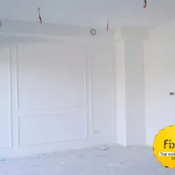 Białe, puste wnętrze po remoncie z dekoracyjnymi panelami ściennymi i widocznymi elementami instalacji elektrycznej w suficie, betonowa podłoga przygotowana do dalszych prac wykończeniowych.