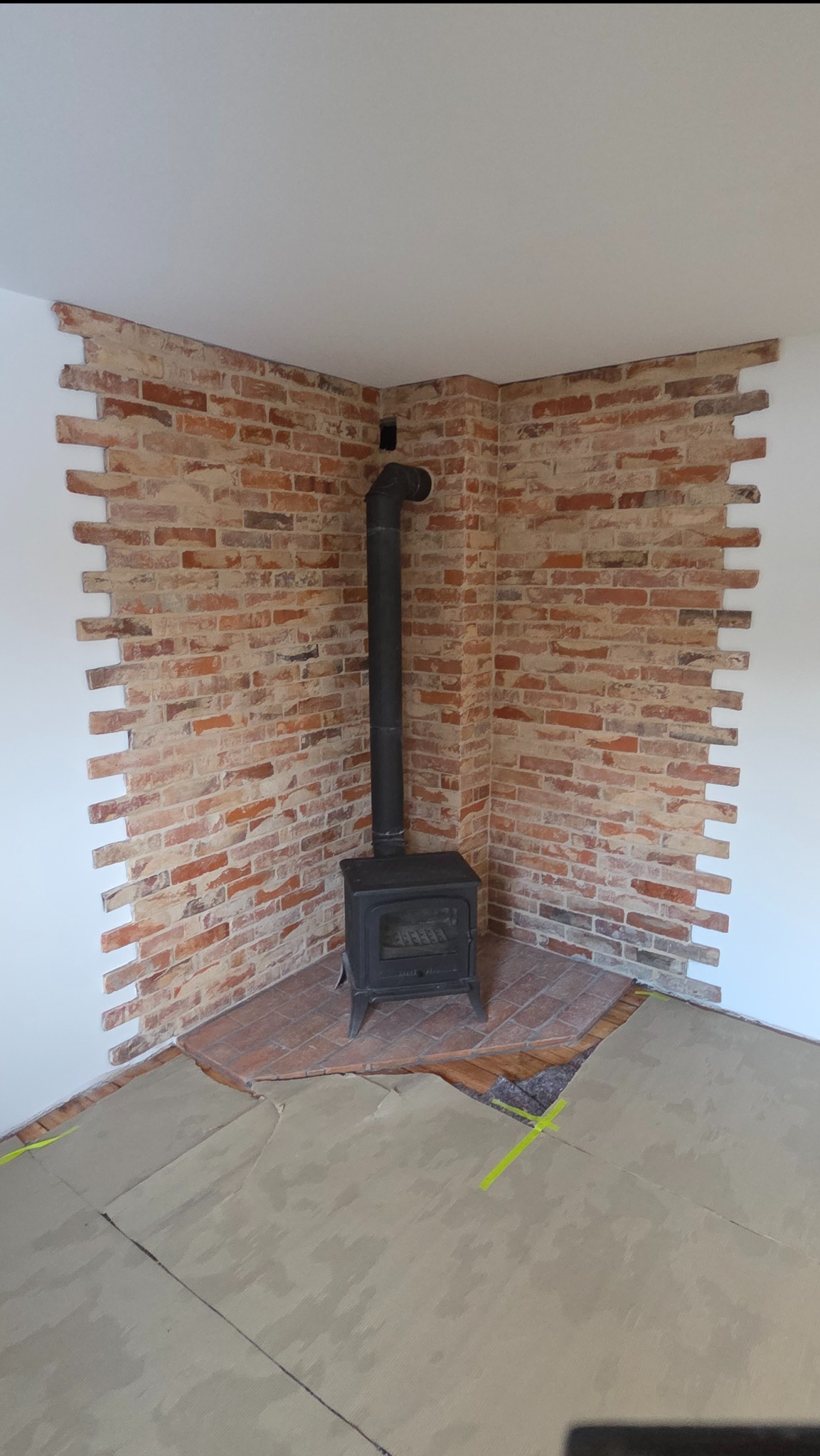Kącik z ceglaną ścianą i piecem kominkowym. Wykończenie ściany z cegły klinkierowej, widoczne zabezpieczenie podłogi przed zabrudzeniem.