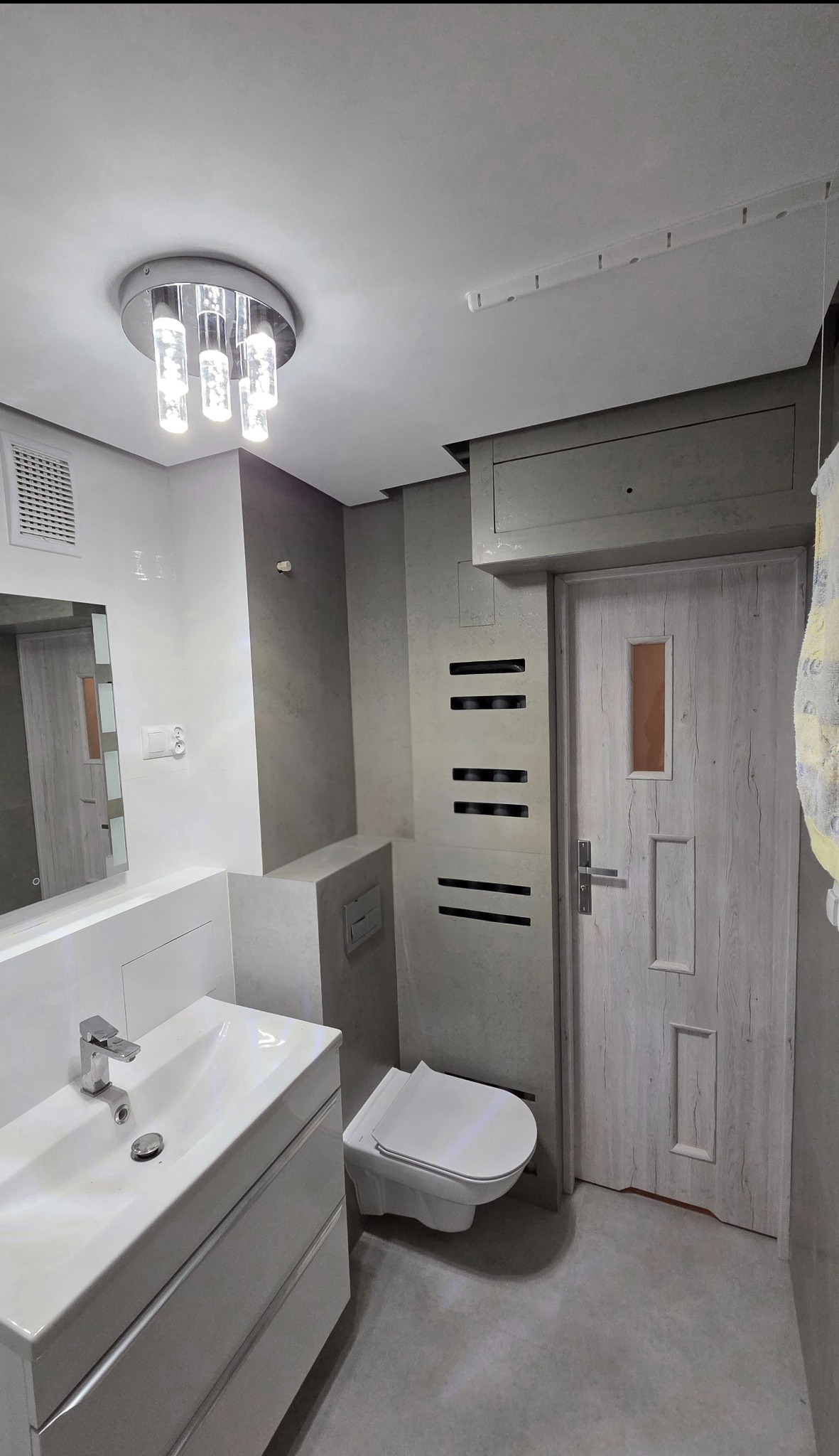 Nowoczesna łazienka w szarościach z podwieszaną toaletą, umywalką z szafką i oświetleniem sufitowym. Wykończenie ścian imitujące beton.