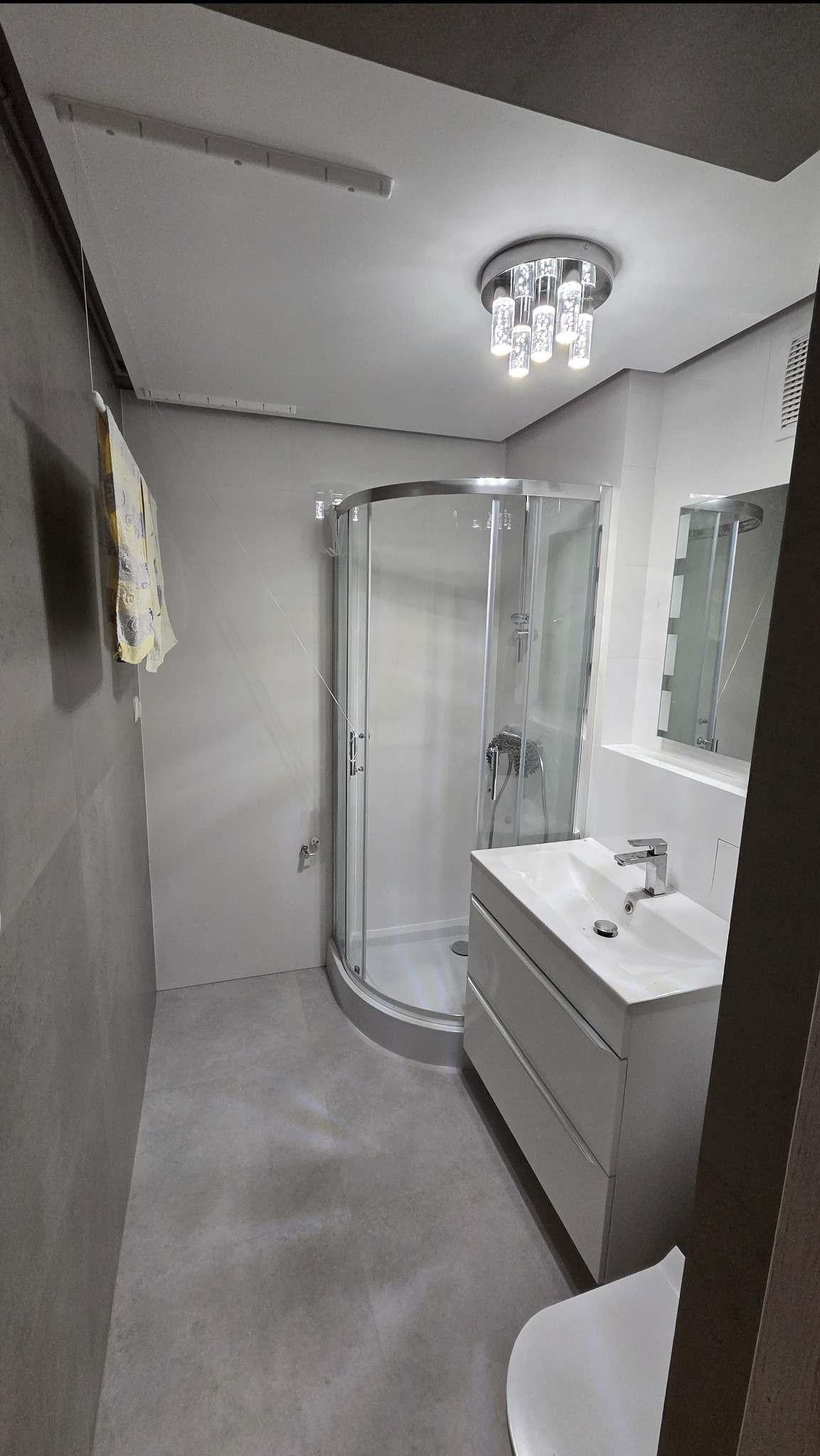 Wykończona łazienka w szaro-białej tonacji z kabiną prysznicową, umywalką z szafką i nowoczesnym oświetleniem sufitowym. Minimalistyczny design.