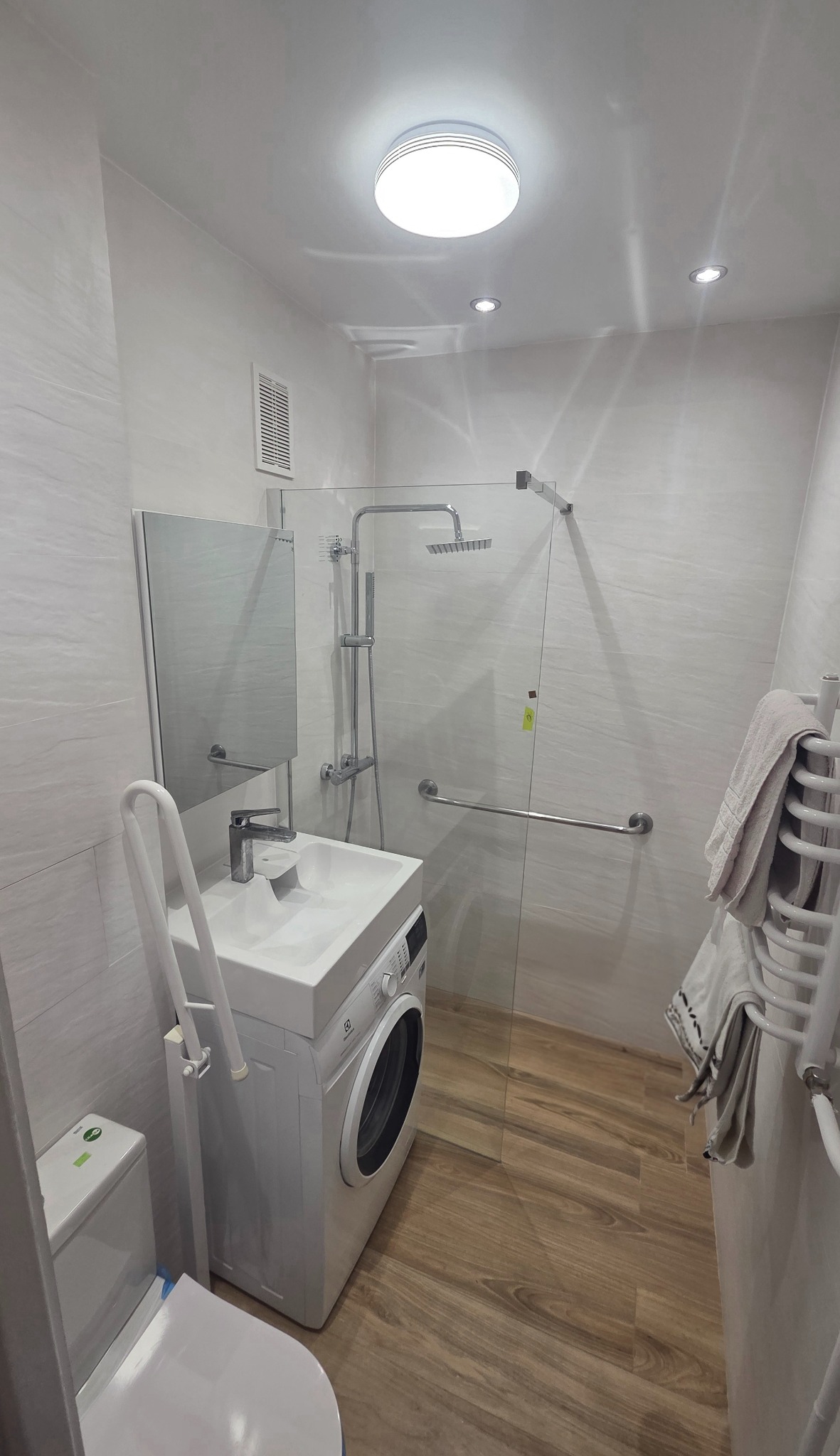 Jasna łazienka po remoncie: kabina prysznicowa ze szklaną ścianką, umywalka z pralką, lustro i drewnopodobna podłoga. Widoczne detale wykończenia.