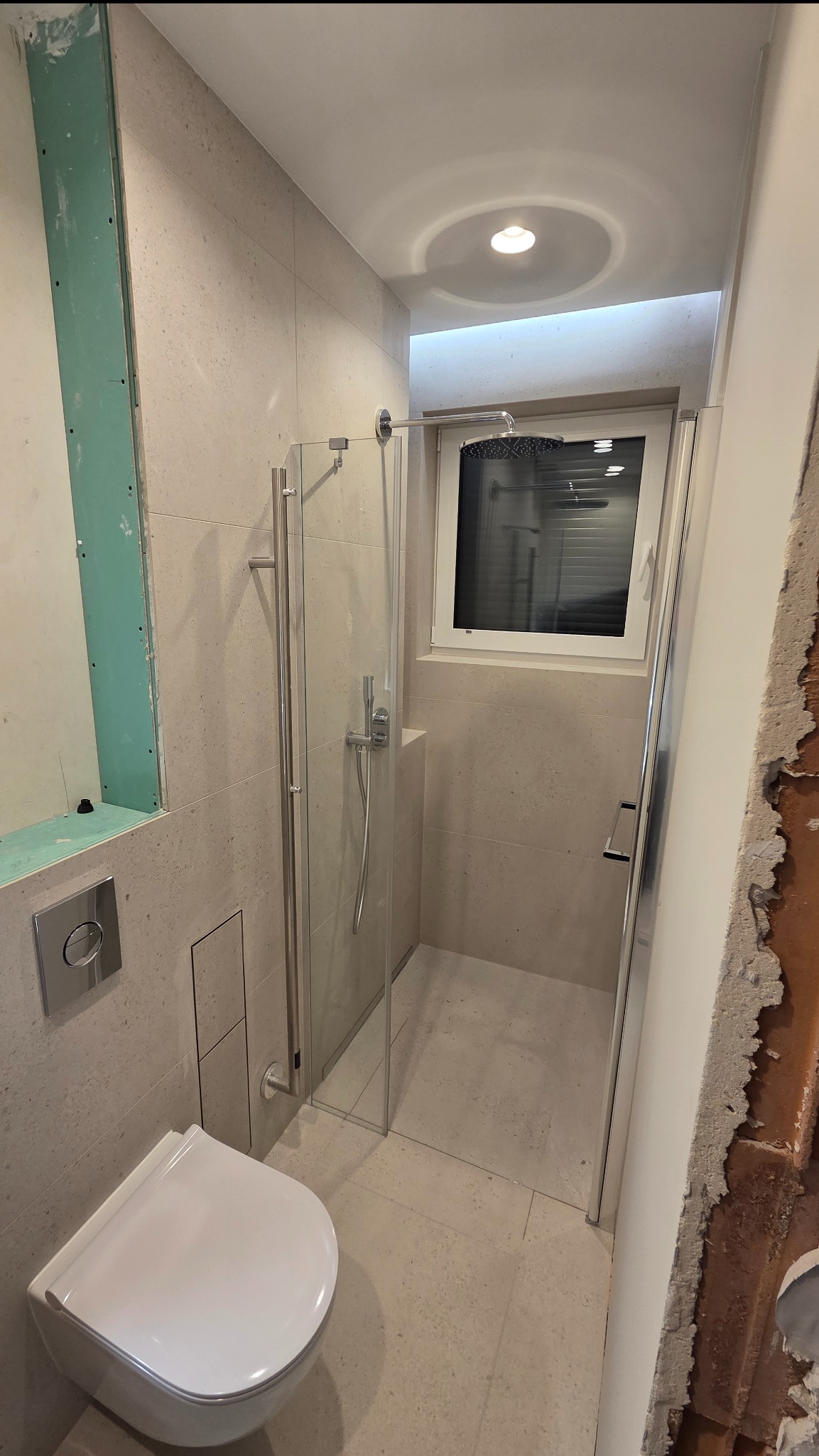 Jasna łazienka z kabiną prysznicową i podwieszaną toaletą. Fragment ściany z płytą gipsową i surowy fragment ściany po prawej stronie. Widoczne okno w kabinie.