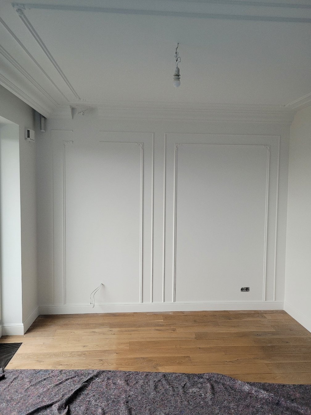 Białe ściany z dekoracyjnymi listwami i drewnianą podłogą. Widoczne gniazdka elektryczne i kabel wystający ze ściany. Na podłodze rozłożona szara mata ochronna.