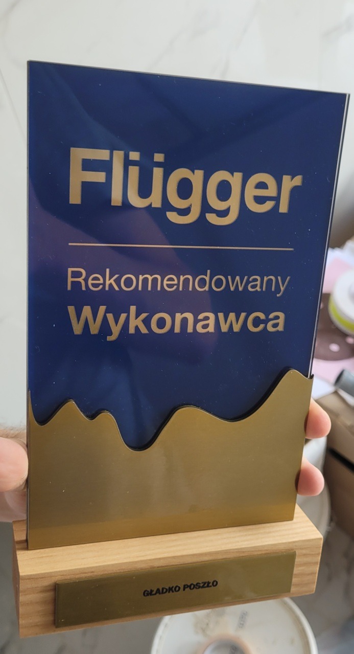 Nagroda 'Rekomendowany Wykonawca Flügger' z napisem 'Gładko Poszło', na drewnianej podstawie, trzymana w dłoni. Niebiesko-złota kolorystyka.