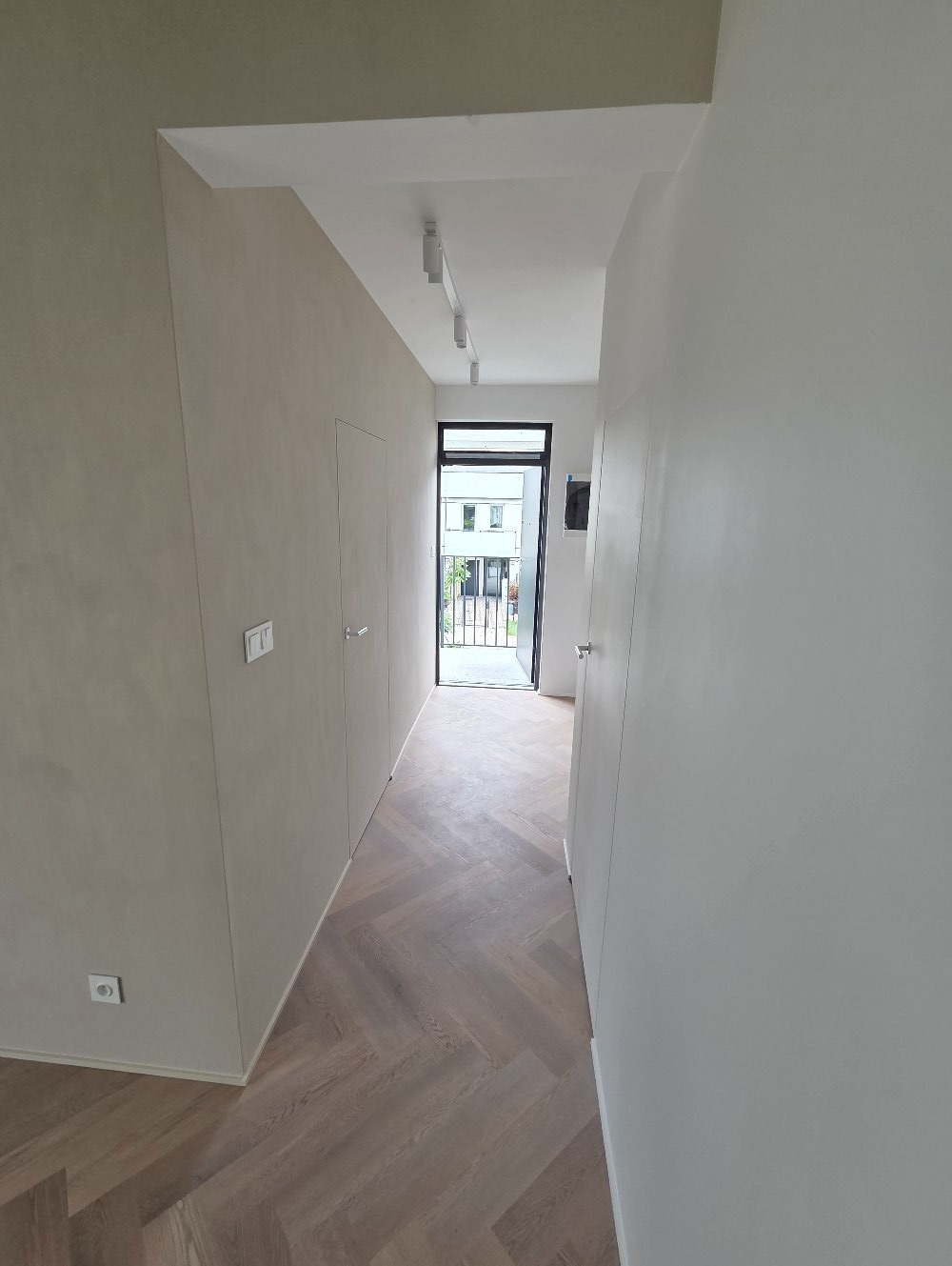 Korytarz po remoncie: ściany w odcieniach beżu i bieli, parkiet ułożony w jodełkę, nowoczesne oświetlenie sufitowe. Widok na zewnątrz przez otwarte drzwi.