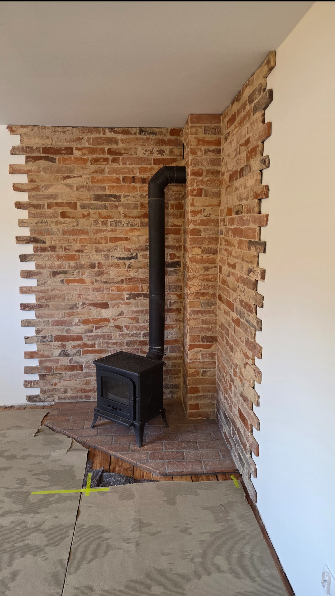 Ceglana ściana z kominkiem w trakcie wykończenia. Fragmenty białych ścian, zabezpieczona podłoga. Widoczne niedokończone łączenia cegieł ze ścianą.