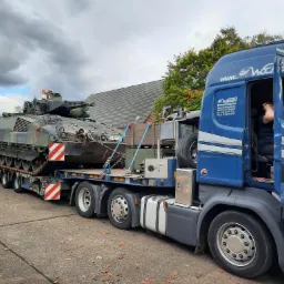 Niebieska ciężarówka z otwartymi drzwiami przewozi wojskowy czołg na niskopodwoziowej przyczepie; widoczne oznaczenia ostrzegawcze na pojeździe, liście na ziemi i budynek w tle.