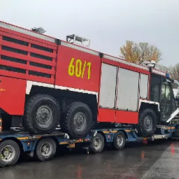 Czerwony wóz strażacki z żółtym napisem 60/1 marki Ziegler transportowany na niskopodwoziowej lawecie, Popowo Kościelne. Na drugim planie inne pojazdy i drzewa.