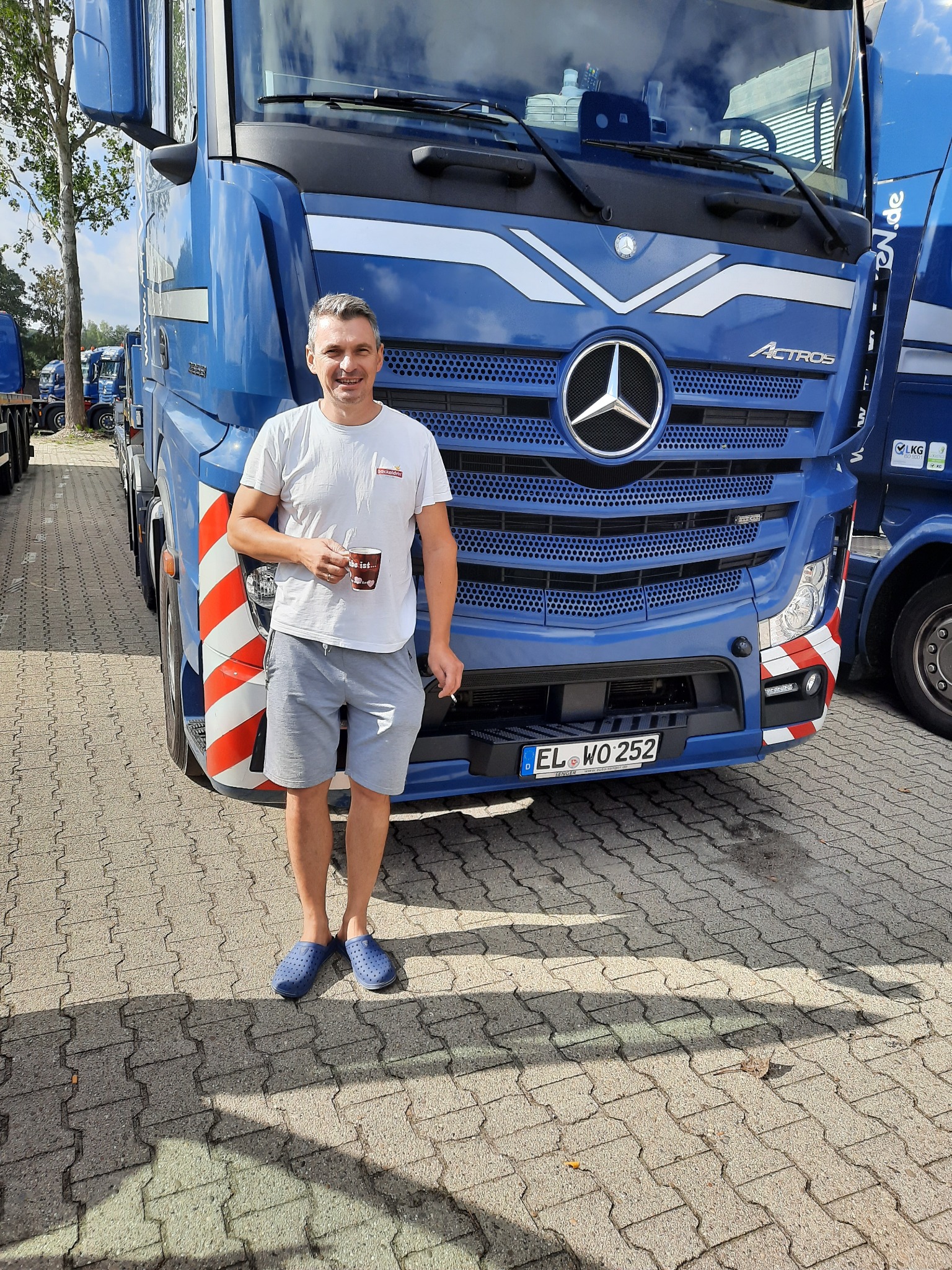 Kierowca z kubkiem stoi przed niebieską ciężarówką Mercedes Actros z niemiecką tablicą rejestracyjną na brukowanym placu.