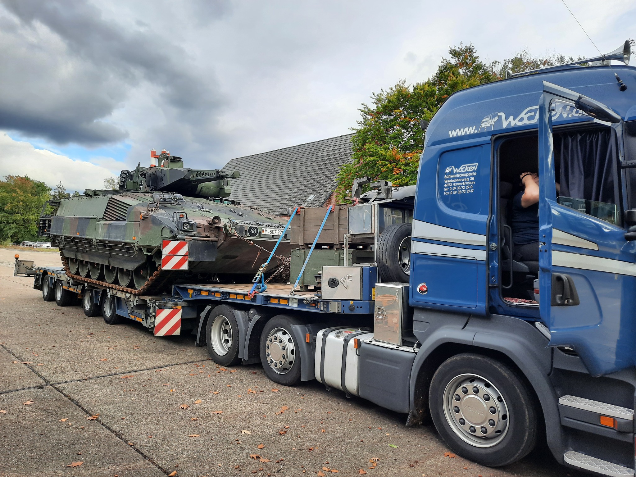 Niebieska ciężarówka z otwartymi drzwiami przewozi wojskowy czołg na niskopodwoziowej przyczepie; widoczne oznaczenia ostrzegawcze na pojeździe, liście na ziemi i budynek w tle.