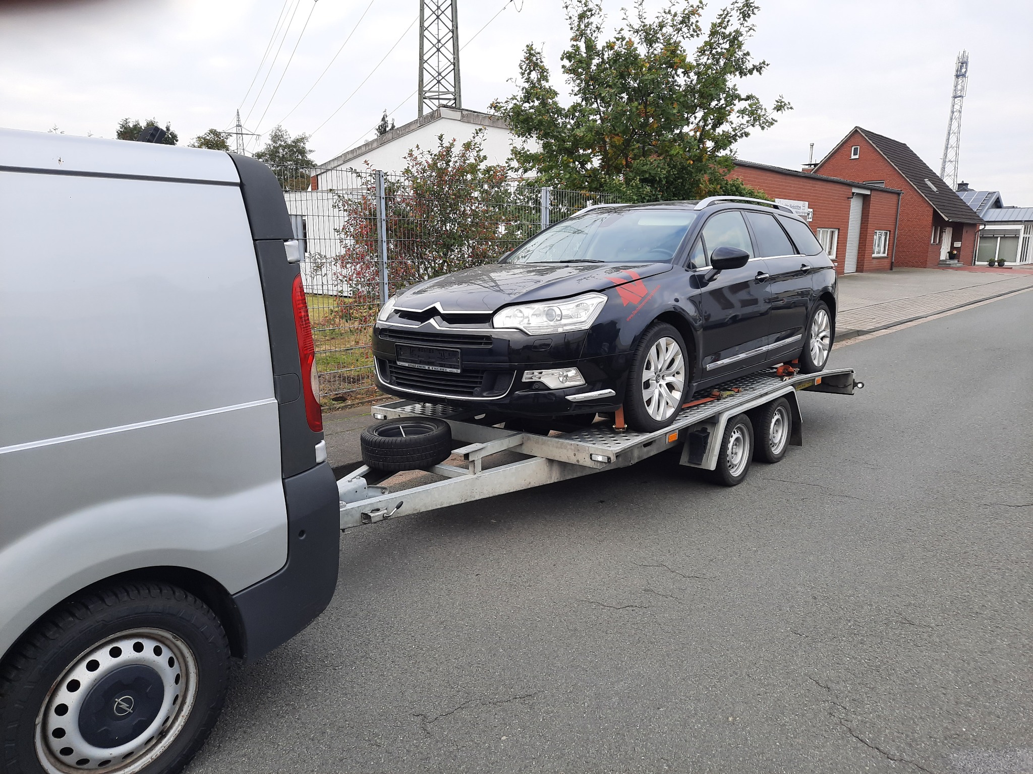 Czarny samochód osobowy Citroen na metalowej lawecie, ciągniętej przez srebrnego busa na tle zabudowań.