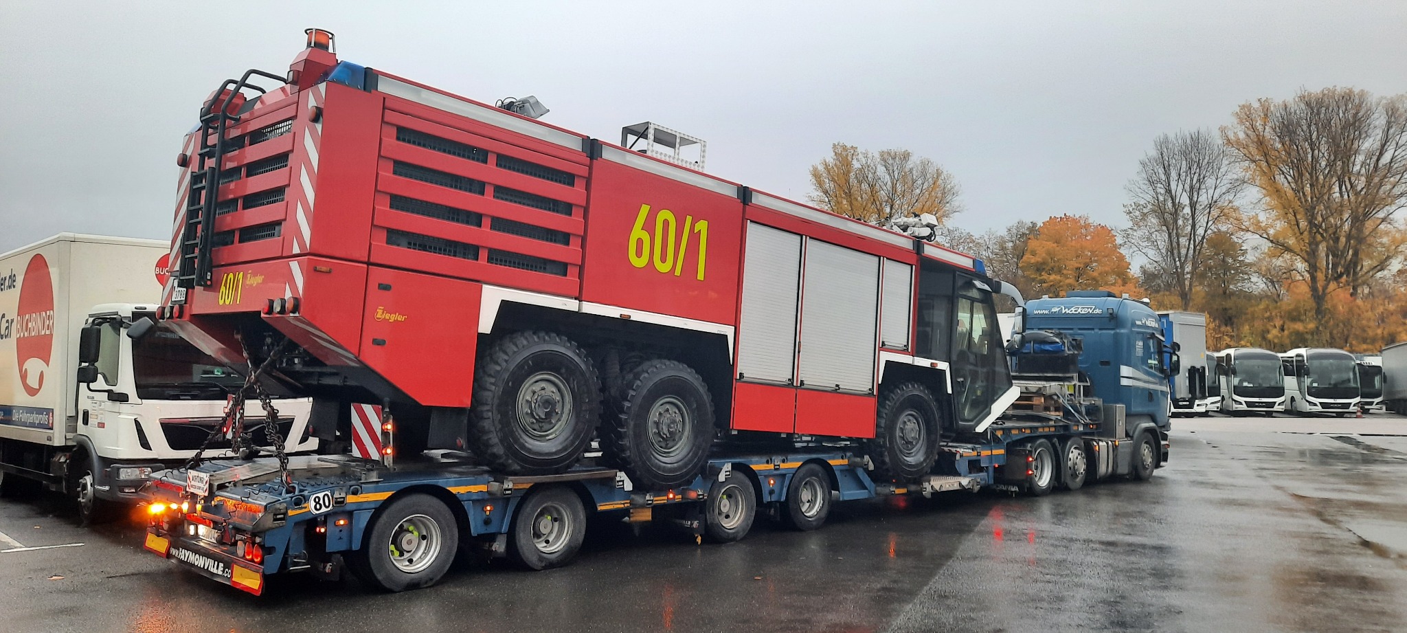 Czerwony wóz strażacki z żółtym napisem 60/1 marki Ziegler transportowany na niskopodwoziowej lawecie, Popowo Kościelne. Na drugim planie inne pojazdy i drzewa.