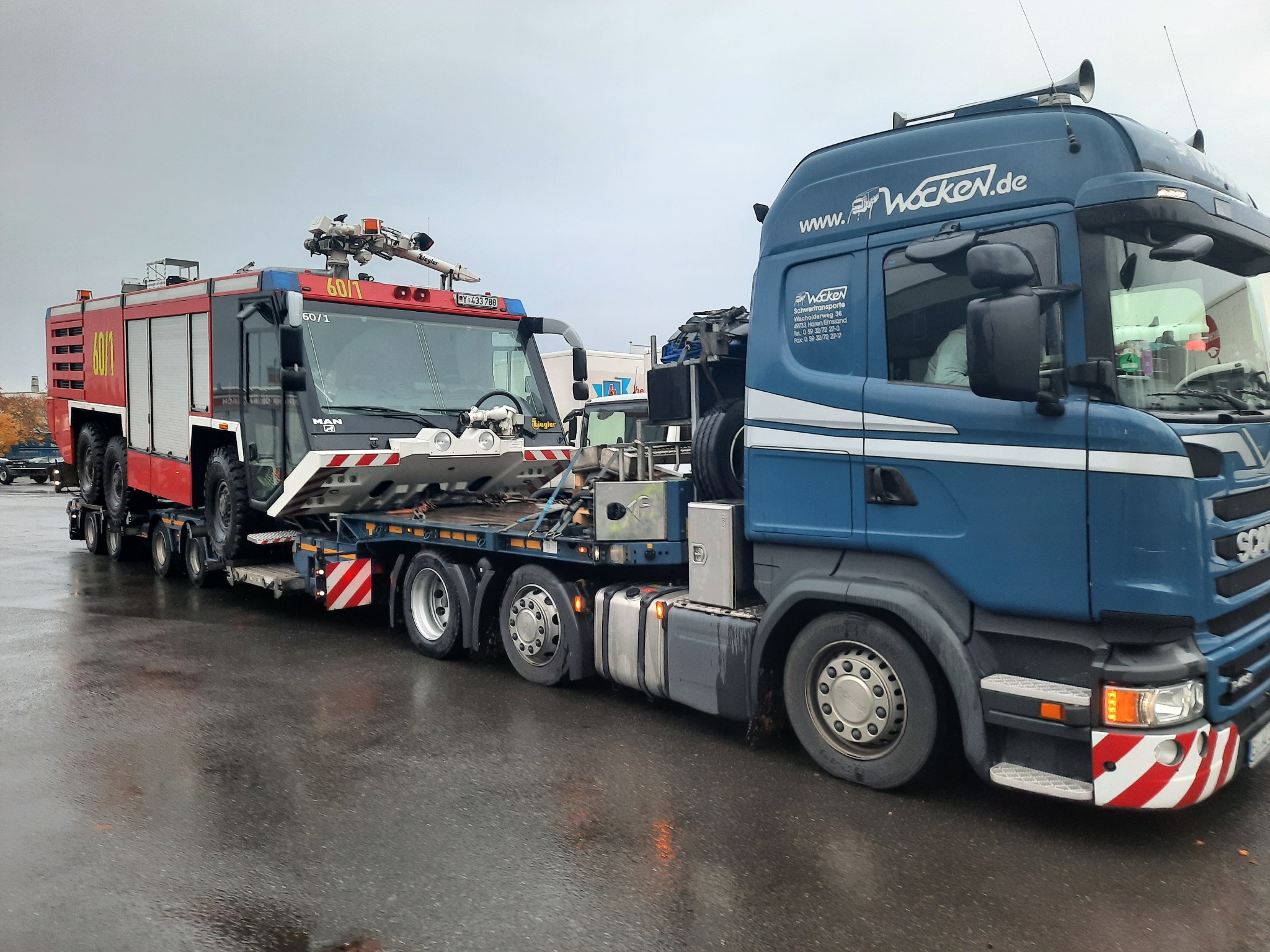 Niebieska ciężarówka Scania z logo firmy Wocken przewozi na niskopodwoziowej lawecie czerwony wóz strażacki MAN Ziegler z numerem 60/1 w pochmurny dzień.