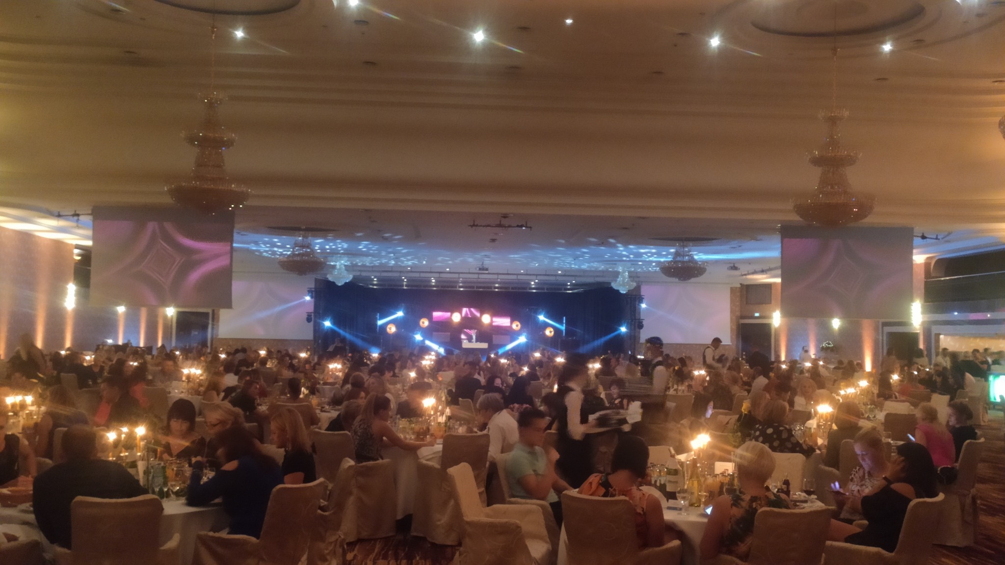 Sala balowa wypełniona gośćmi siedzącymi przy okrągłych stołach, oświetlona świecami i reflektorami sceny z ekranami po bokach, obsługa w tle.