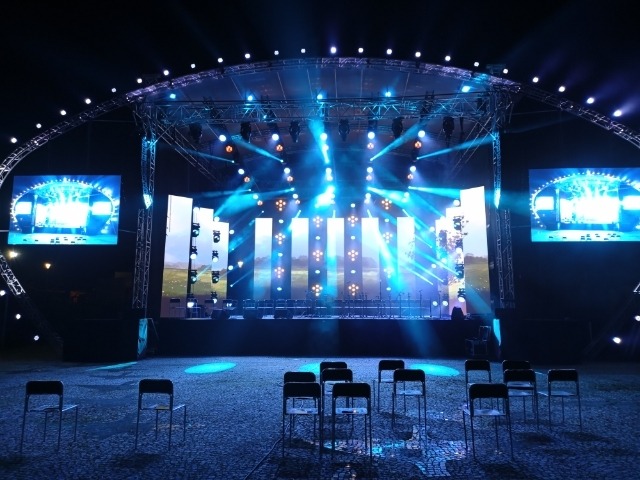 Scena plenerowa z oświetleniem punktowym i ekranami LED, przygotowana do koncertu, z rzędem pustych krzeseł na pierwszym planie.