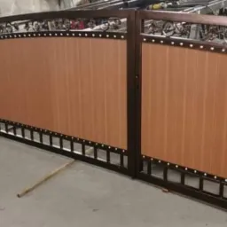 Dwuskrzydłowa brama wjazdowa z brązowymi panelami i dekoracyjnymi elementami metalowymi, ustawiona na drewnianych podporach w hali produkcyjnej.