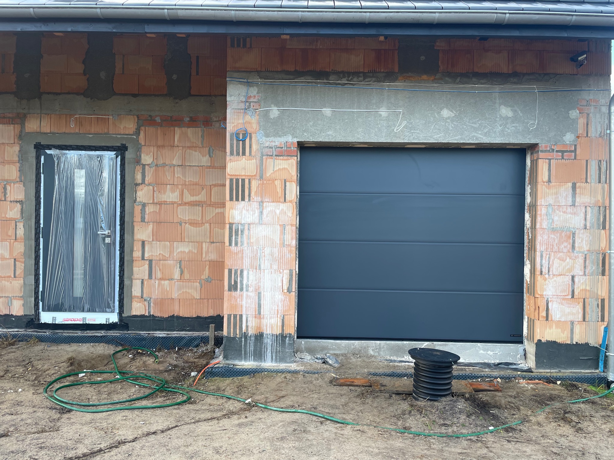 Szara brama garażowa segmentowa w surowym stanie, widoczna instalacja elektryczna, budynek w trakcie budowy, cegły ceramiczne, folia ochronna na oknie.