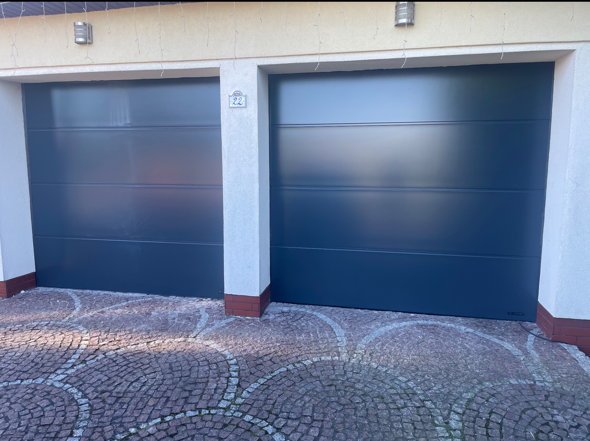 Dwie nowoczesne, grafitowe bramy garażowe segmentowe z poziomymi przetłoczeniami, zamontowane w jasnym budynku z numerem 22, na tle brukowanej nawierzchni.
