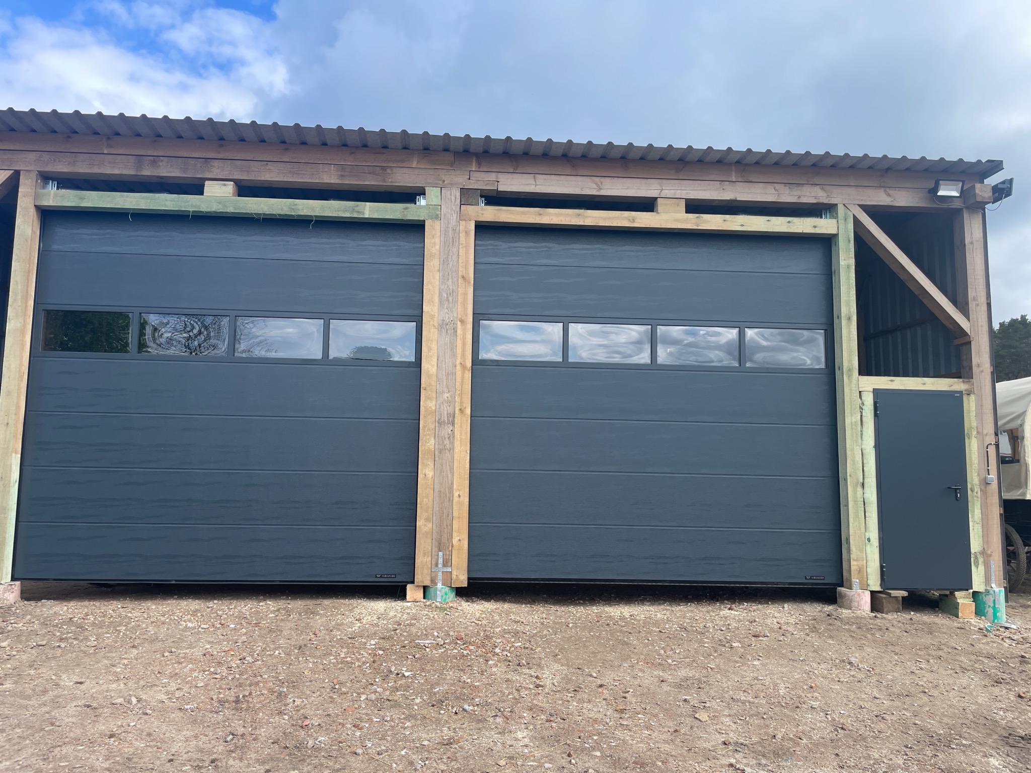 Dwie szare bramy garażowe segmentowe z oknami, zamontowane w drewnianej konstrukcji z blachą falistą na dachu, widoczna mała furtka po prawej stronie, teren wokół bramy utwardzony żwirem.