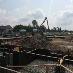 Wykop pod fundamenty z szalunkiem i zbrojeniem, widoczny betonowóz z pompą do betonu oraz hałda ziemi, w tle budynek z napisem Studio Mebli.
