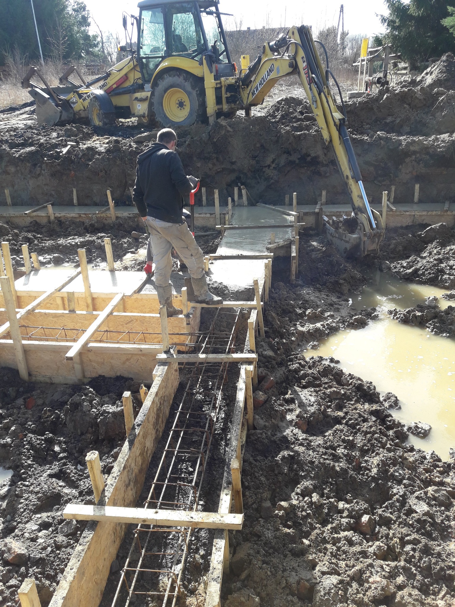 Prace ziemne i wylewanie fundamentów w Lubaniu: widoczny szalunek zbrojeniowy, koparka New Holland i pracownik z łopatą na budowie.