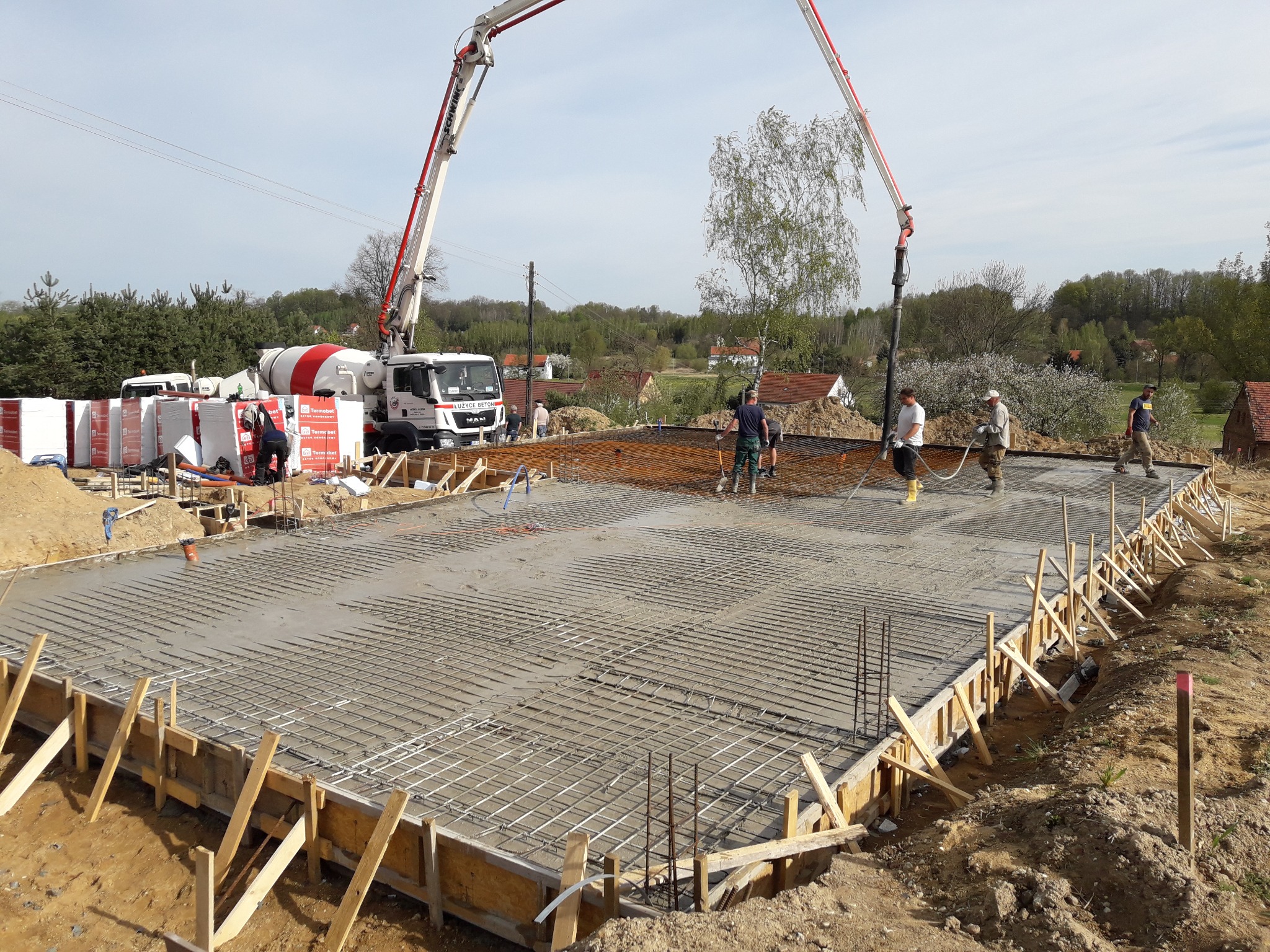 Wylewanie betonu na szalunek zbrojonego fundamentu domu jednorodzinnego, widoczny betonowóz i pracownicy w trakcie pracy.