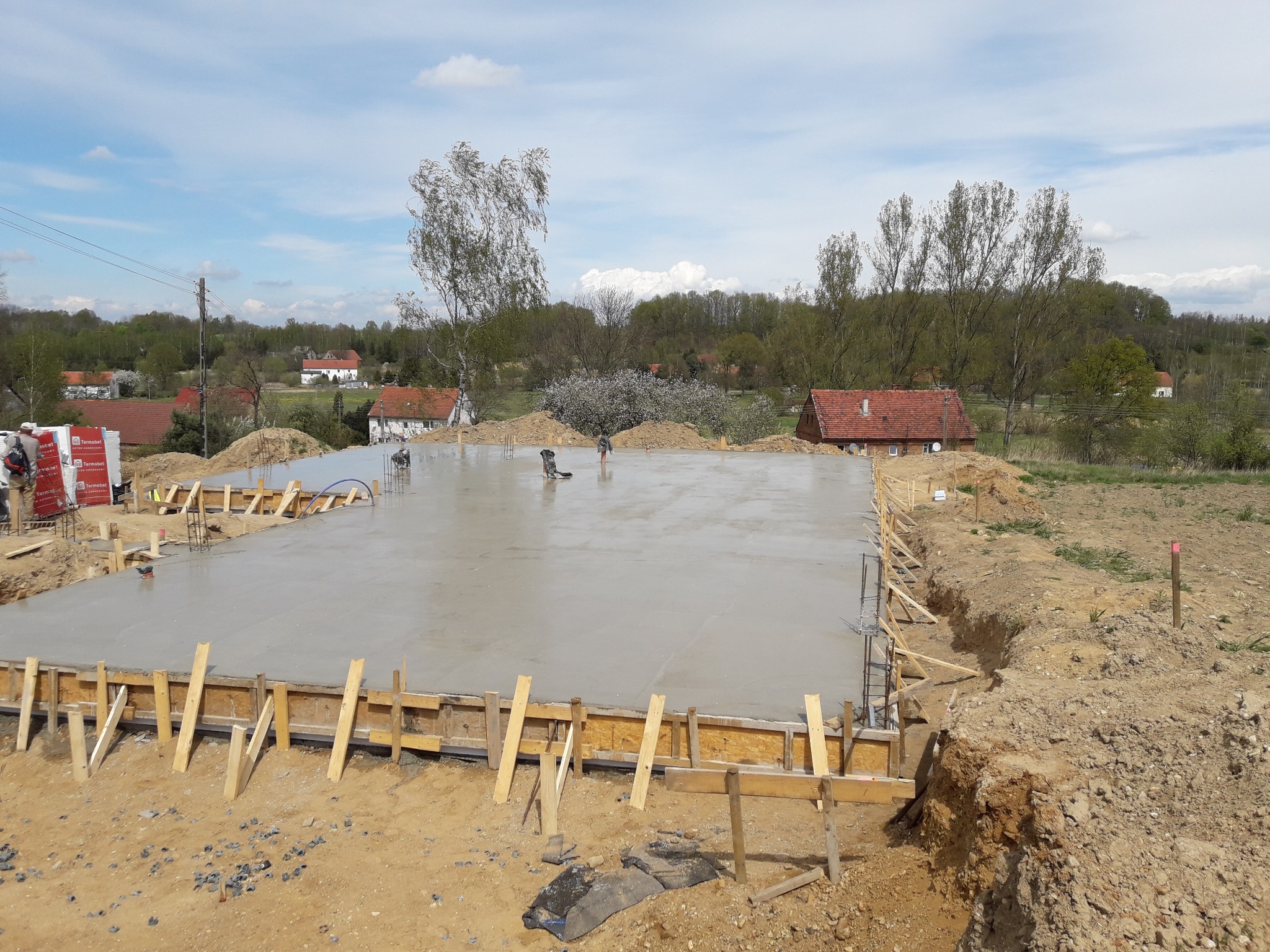 Wylany, świeży fundament płyty betonowej z drewnianym szalunkiem, widoczne zbrojenia i pracownicy w oddali, wiosenny krajobraz w tle.