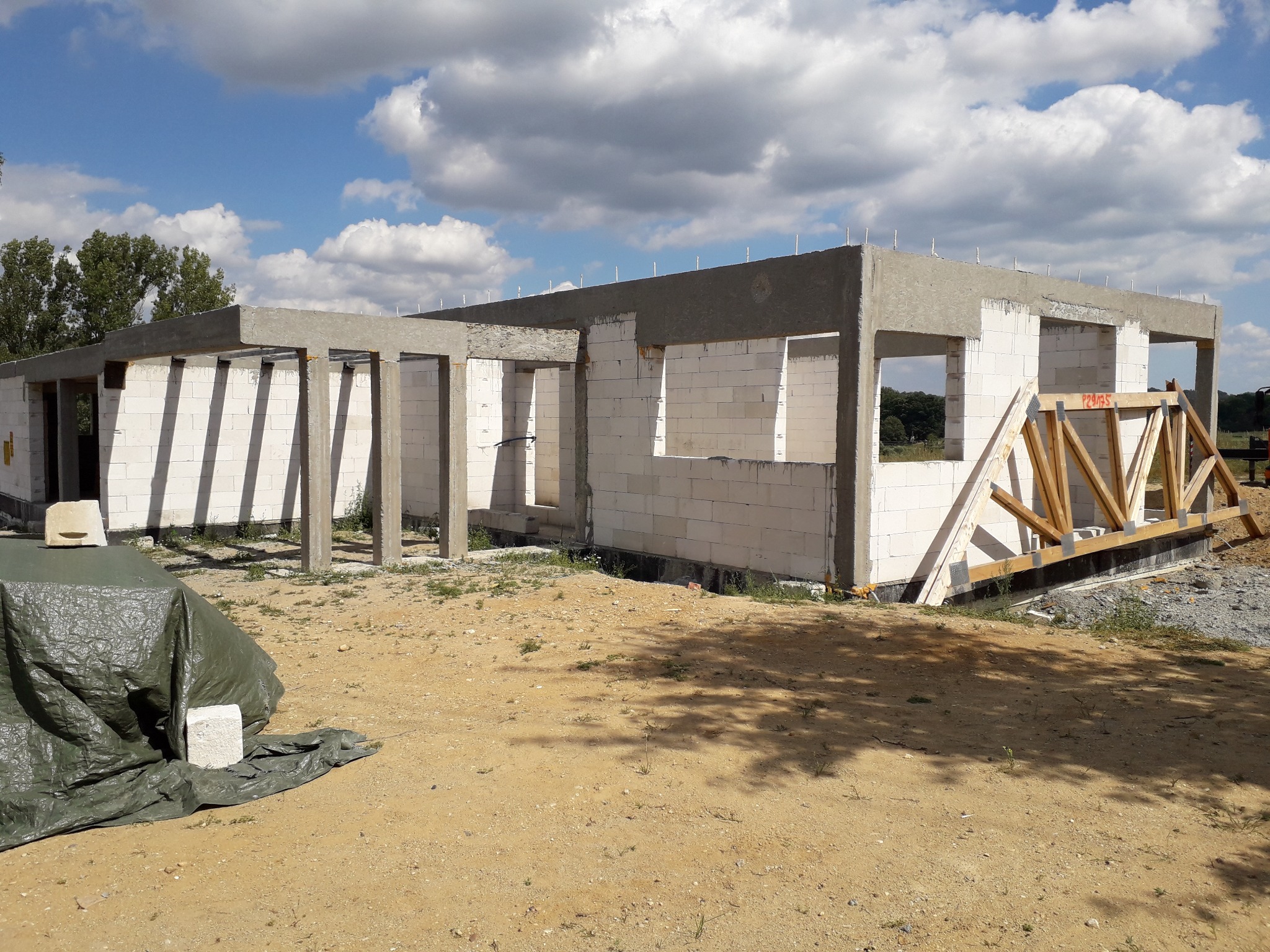 Budowa domu w stanie surowym otwartym z bloczków, widoczne zbrojenia pionowe, drewniane elementy konstrukcji dachowej, teren budowy z piaskiem i częściowo zakryty plandeką.