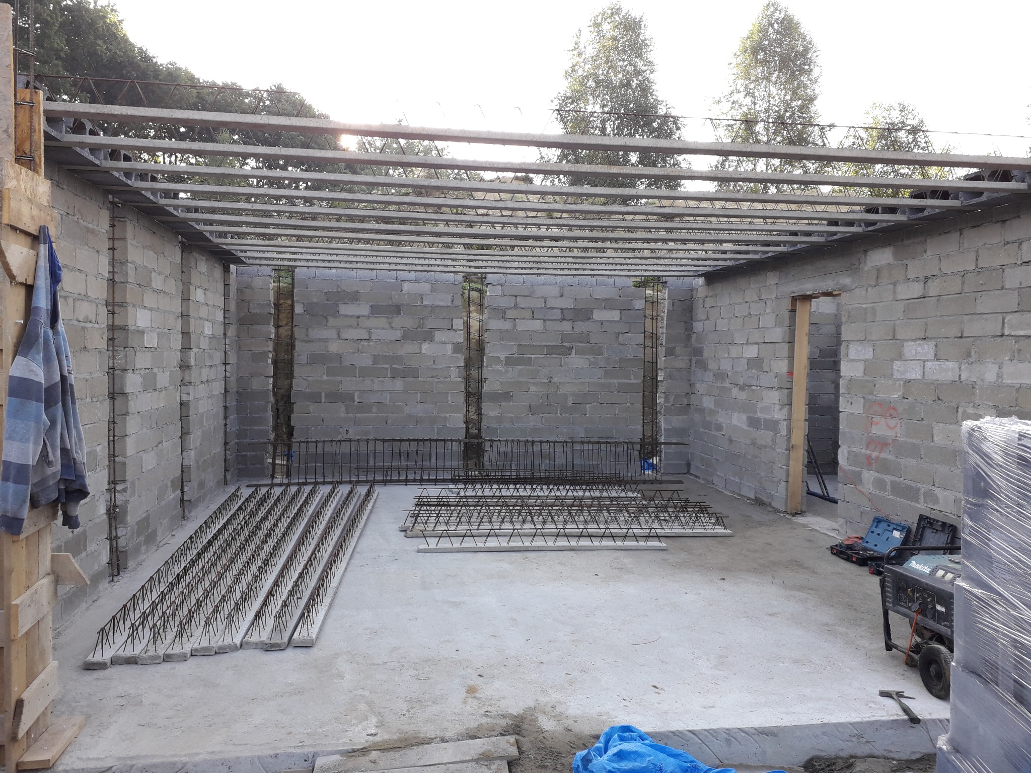 Wnętrze surowego budynku z bloczków betonowych z częściowo wykonanym stropem żelbetowym; widoczne zbrojenia stropowe na podłodze i generator prądu.