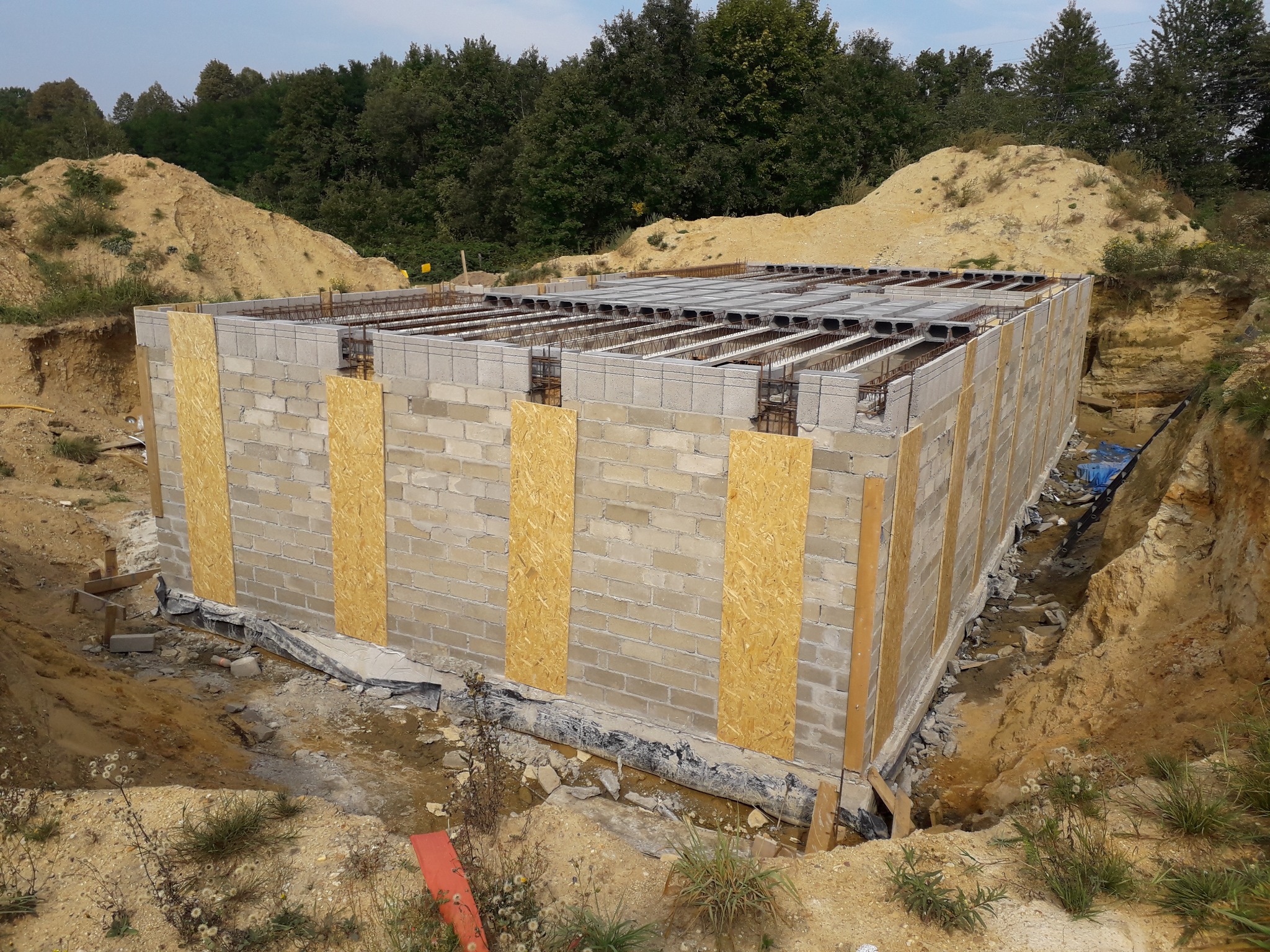 Fundamenty z bloczków betonowych, zabezpieczone płytami OSB, z widocznym zbrojeniem stropu, w wykopie z hałdami ziemi w tle.