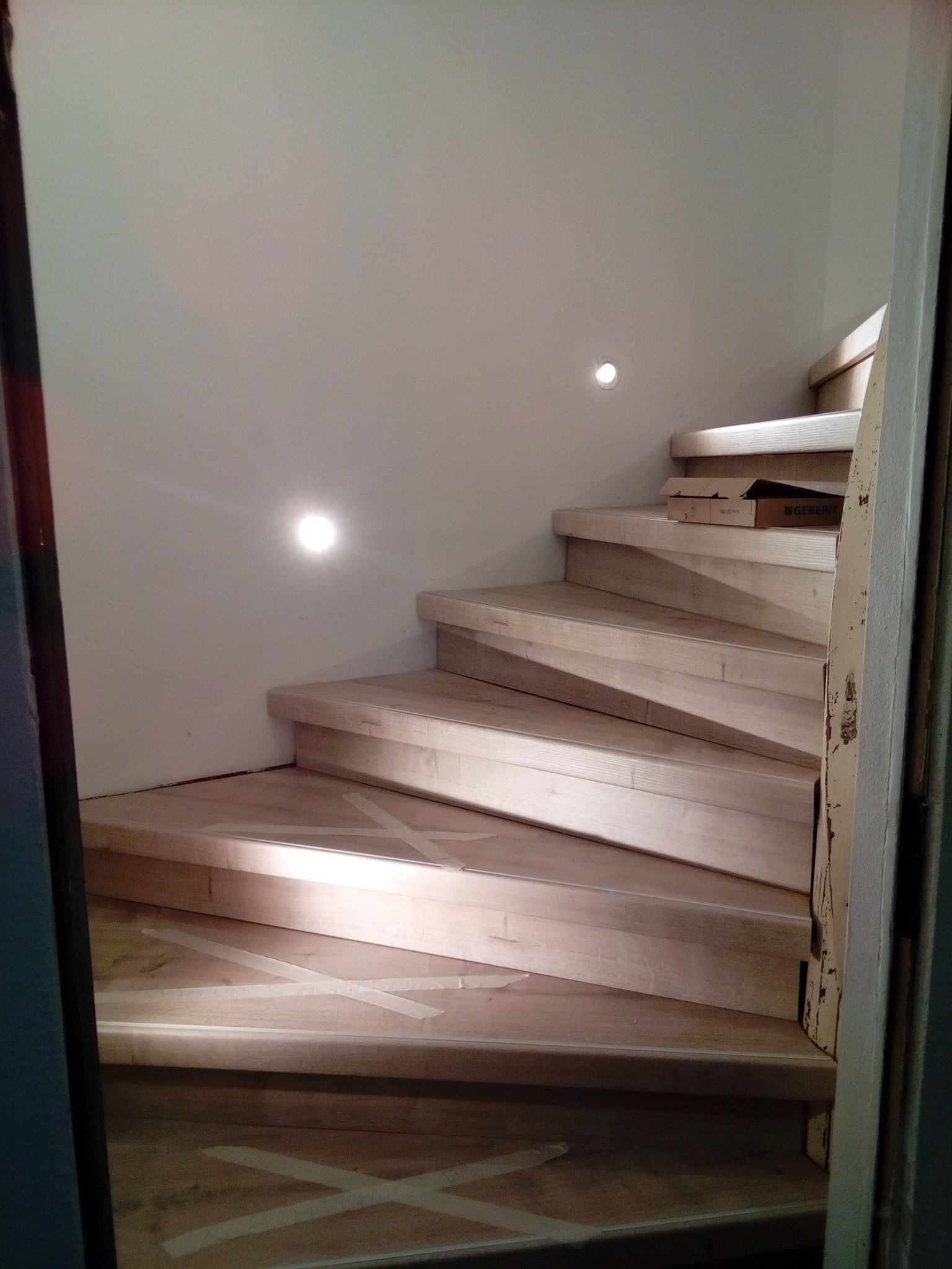 Drewniane schody spiralne w trakcie montażu, zabezpieczone taśmą malarską, z oświetleniem punktowym w ścianie i kartonem Geberit na jednym ze stopni.