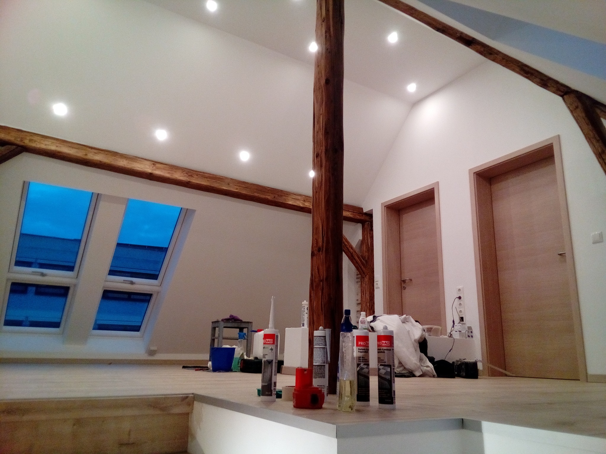 Poddasze w trakcie prac wykończeniowych: widoczne okna dachowe, drewniana kolumna konstrukcyjna, drzwi wewnętrzne oraz tuby z silikonem i akrylem na pierwszym planie.
