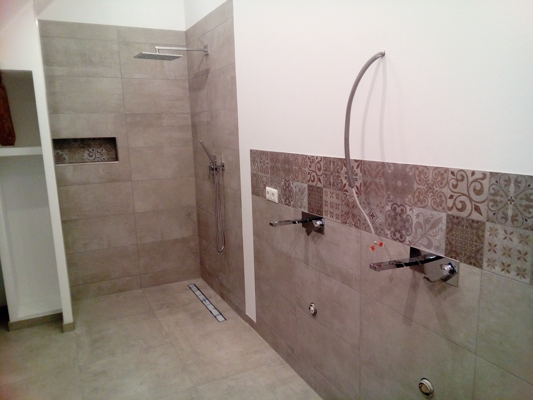 Łazienka w trakcie wykańczania: szare płytki, chromowana armatura, dekoracyjny pas płytek z wzorami, widoczny odpływ liniowy w podłodze i wystające przewody instalacyjne.