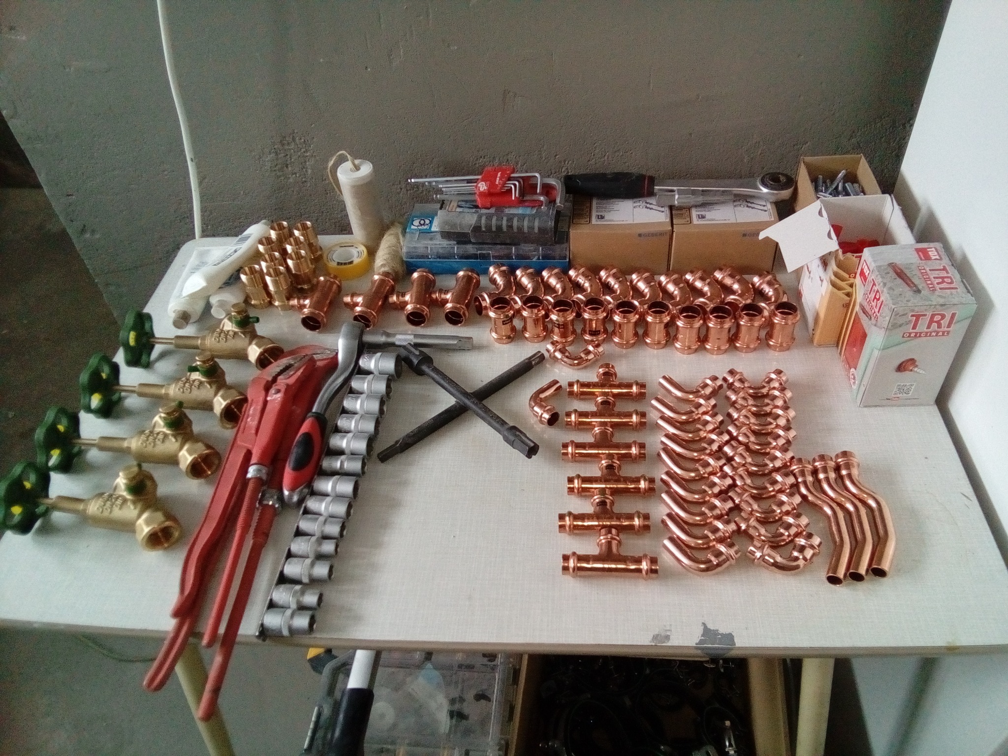 Miedziane kształtki hydrauliczne, zawory z zielonymi pokrętłami i narzędzia na białym stole warsztatowym w pomieszczeniu.