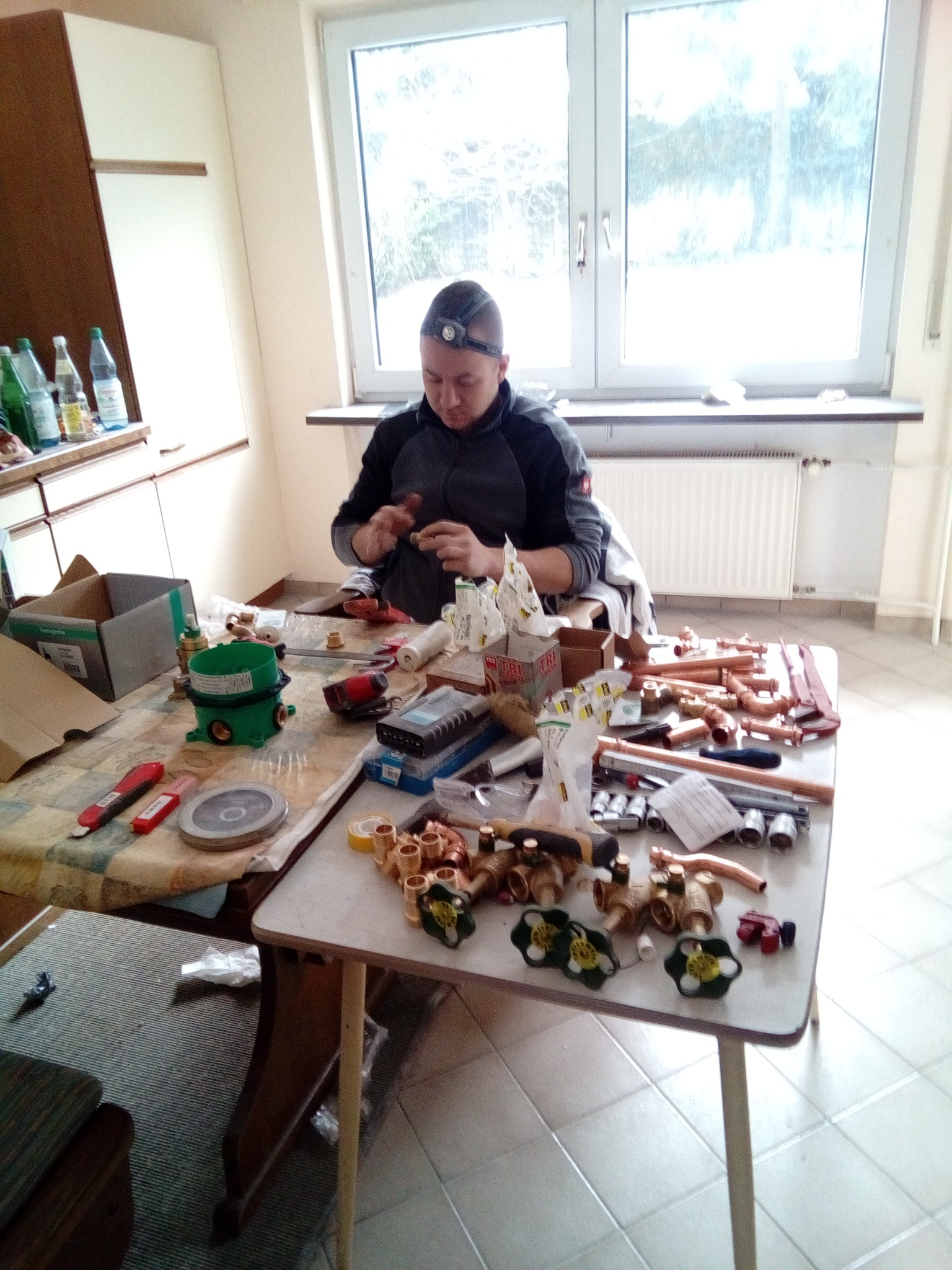 Hydraulik łączy miedziane rury i zawory na stole w jasnym pomieszczeniu, używając narzędzi i oświetlenia czołowego.